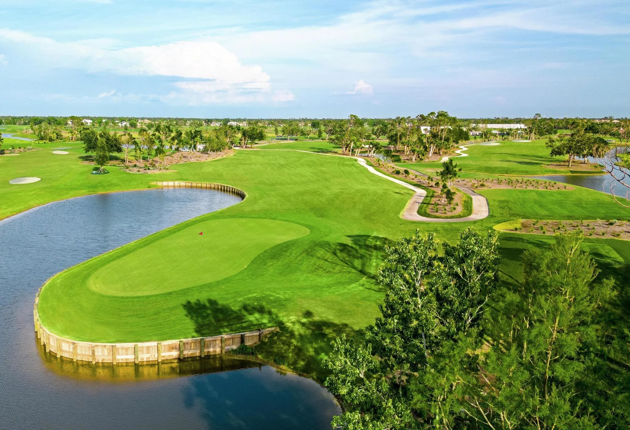 Aerial view of a golf course with lush green grass, water hazards, sand bunkers, and scattered trees under a partly cloudy sky.