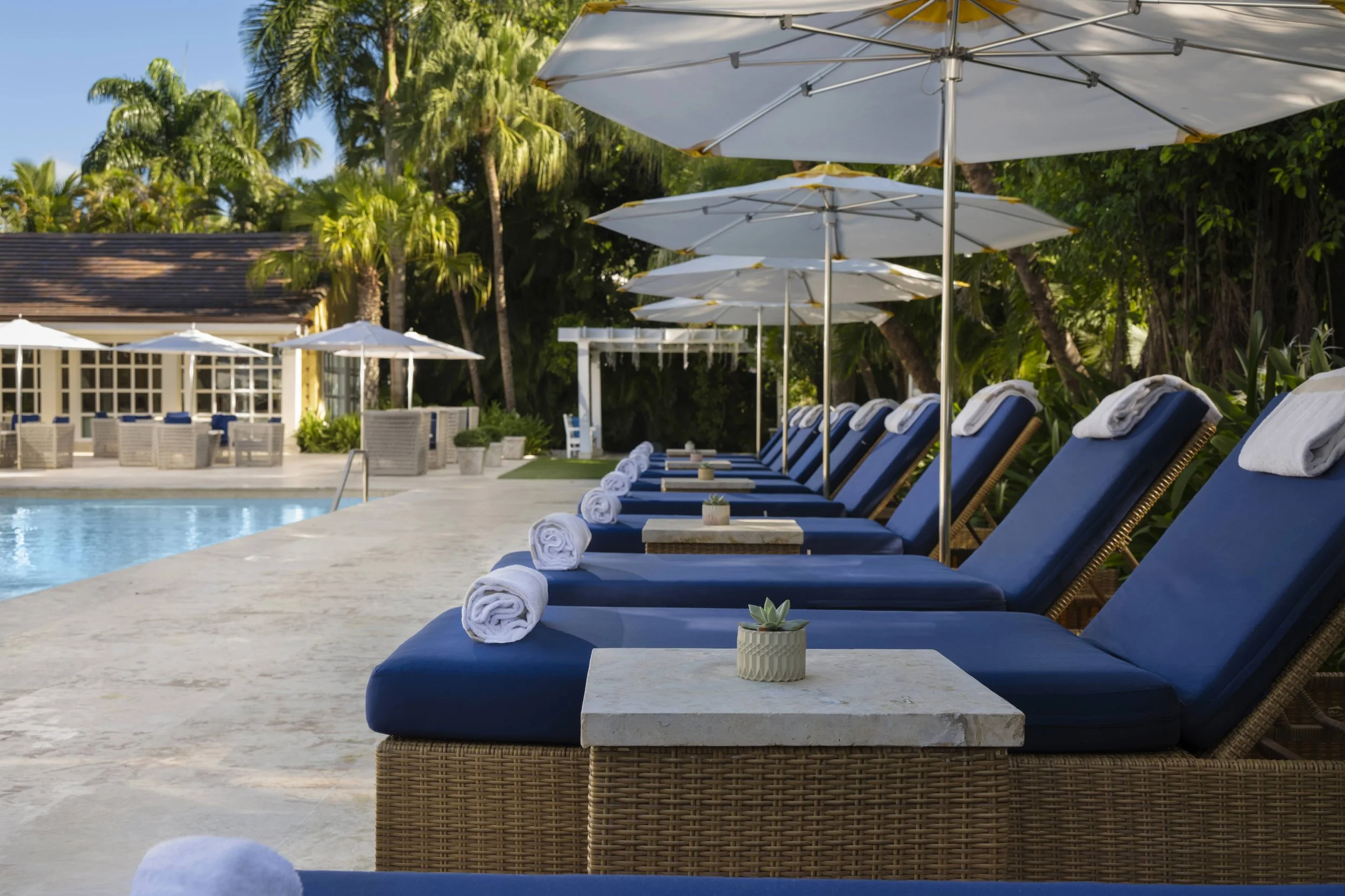 Poolside lounge chairs with rolled towels and small tables under white umbrellas near a swimming pool, with palm trees and a building in the background.