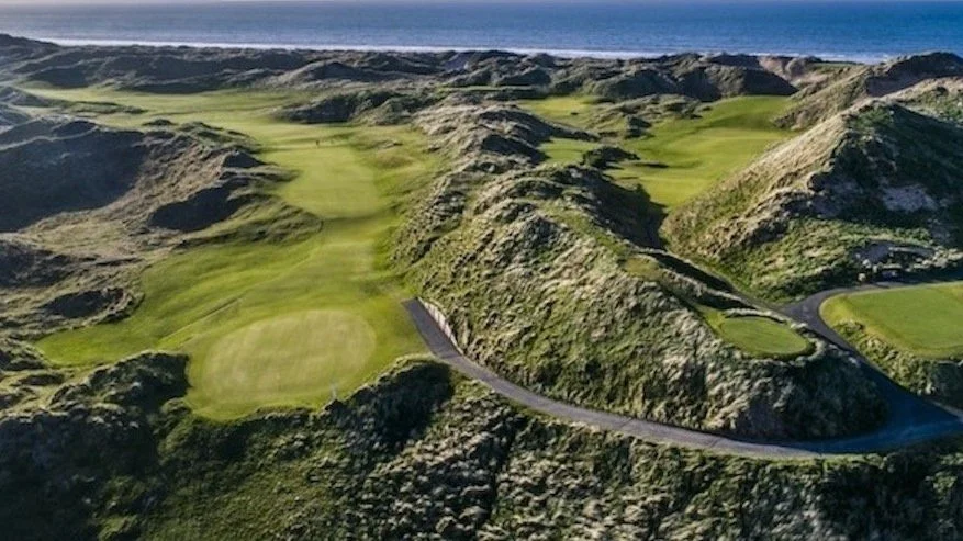 Aerial view of a golf course with rolling green hills and valleys, bordered by a coastal ocean under a clear blue sky.