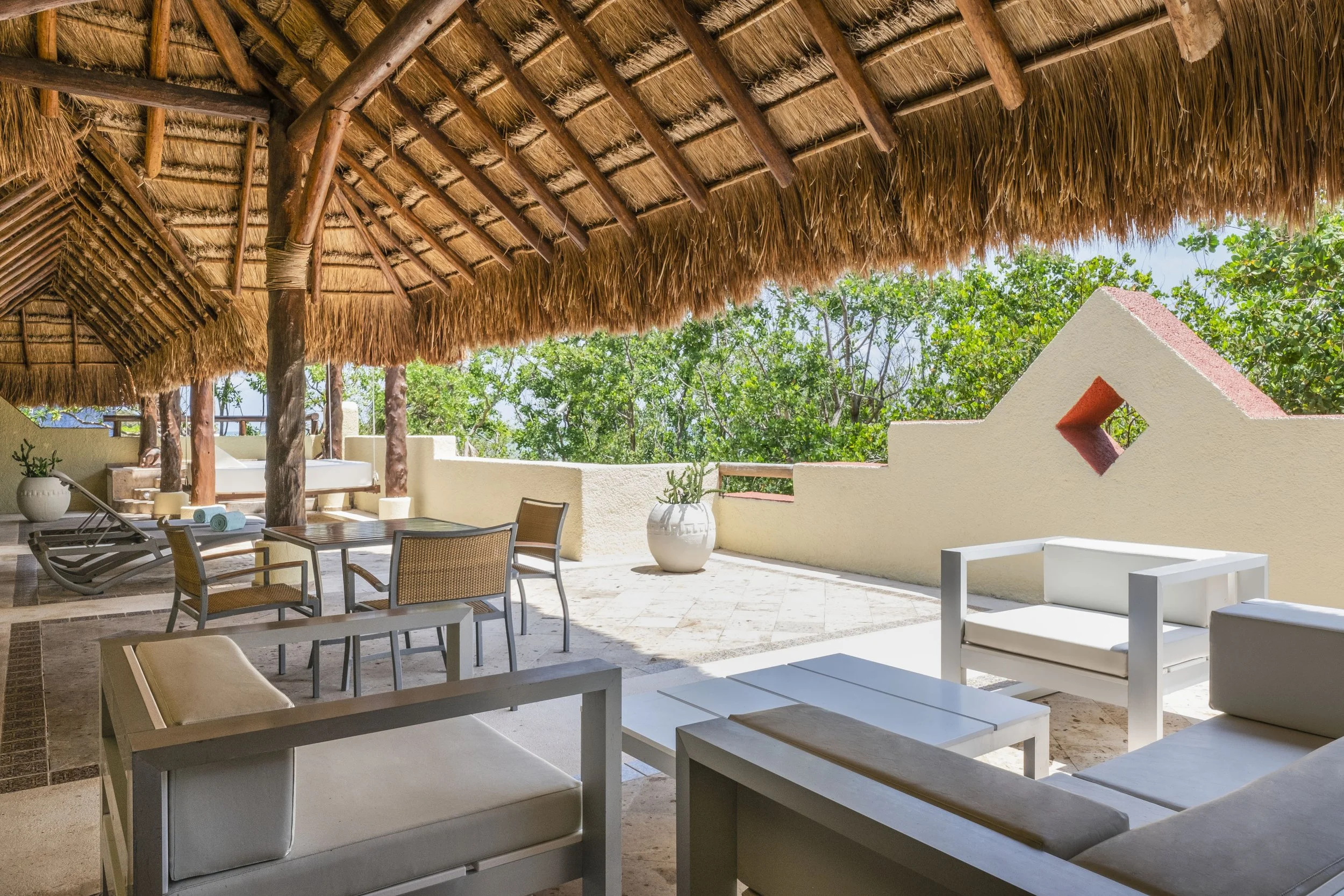 Open-air terrace with thatched roof, white outdoor furniture, potted plants, and greenery in the background.