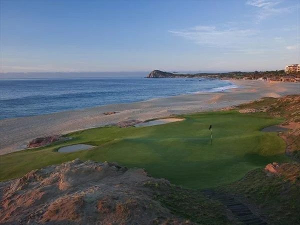 Golf course with green grass, flagstick, and small water hazards on a coastal beach with ocean and rocky shoreline in the background.