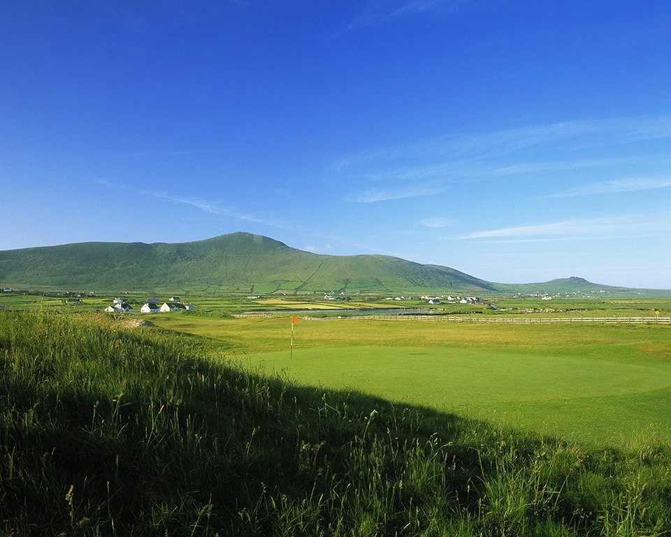 Scenic view of a golf course with a green grass putting area, a flagstick, and rolling hills with small houses under a blue sky with wispy clouds.