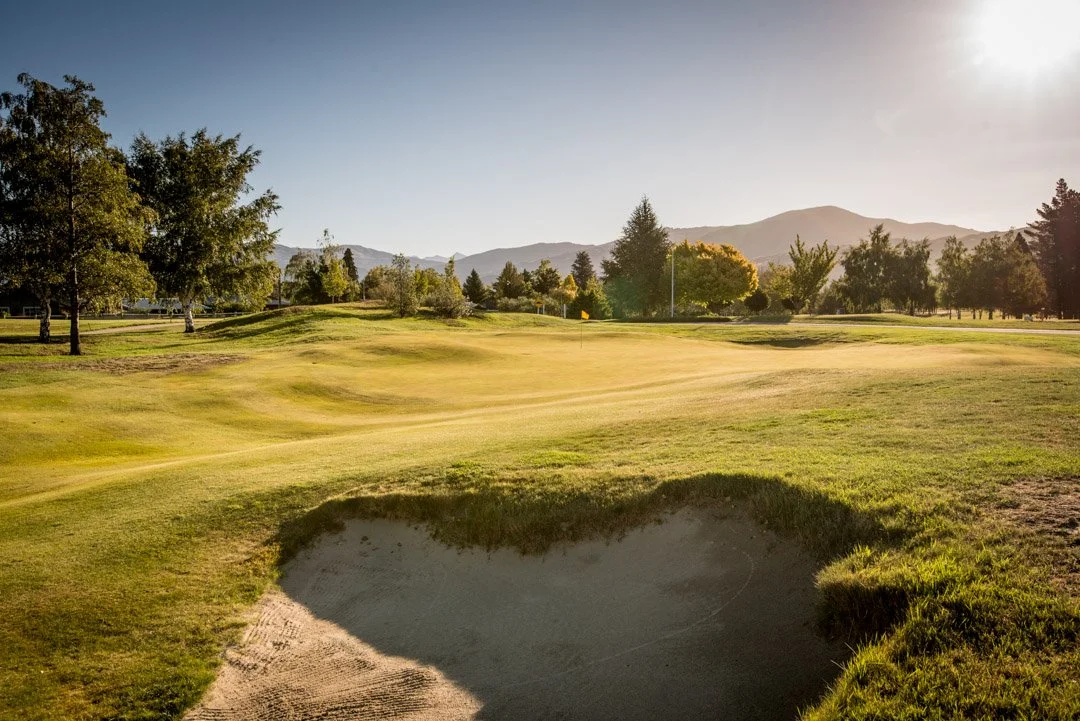 Golf course with a sand trap, green grass, trees, and mountains in the background during sunset.