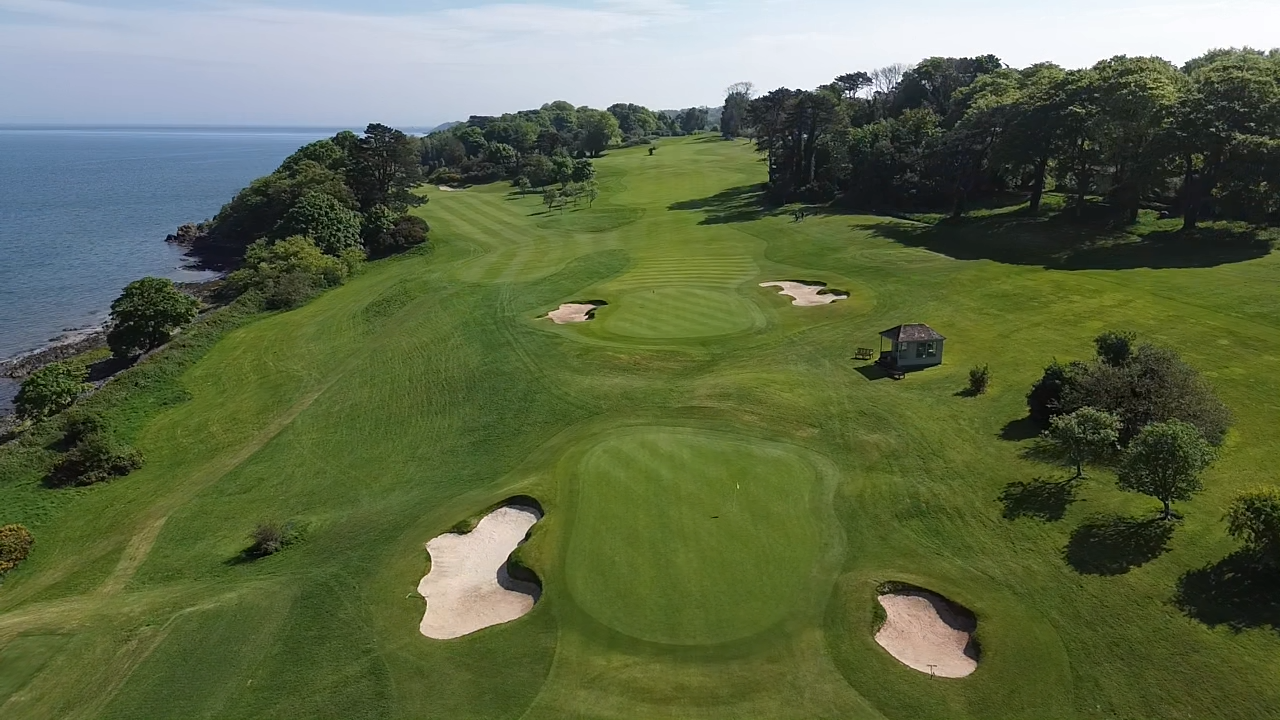 An aerial view of a golf course located by the water, featuring well-maintained greens, sand bunkers, scattered trees, and a small building on the right side.