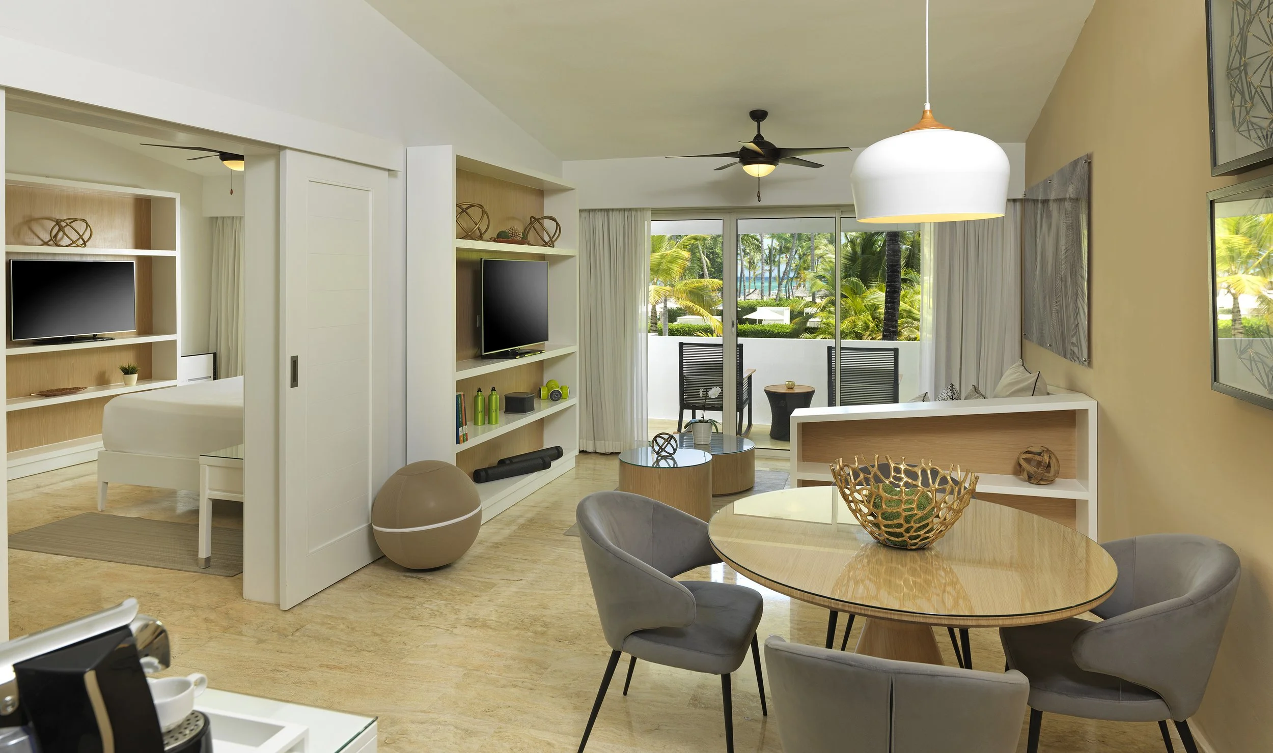 Living room with a round dining table, gray chairs, a TV on a shelf, sliding door leading to a balcony with chairs and trees outside.