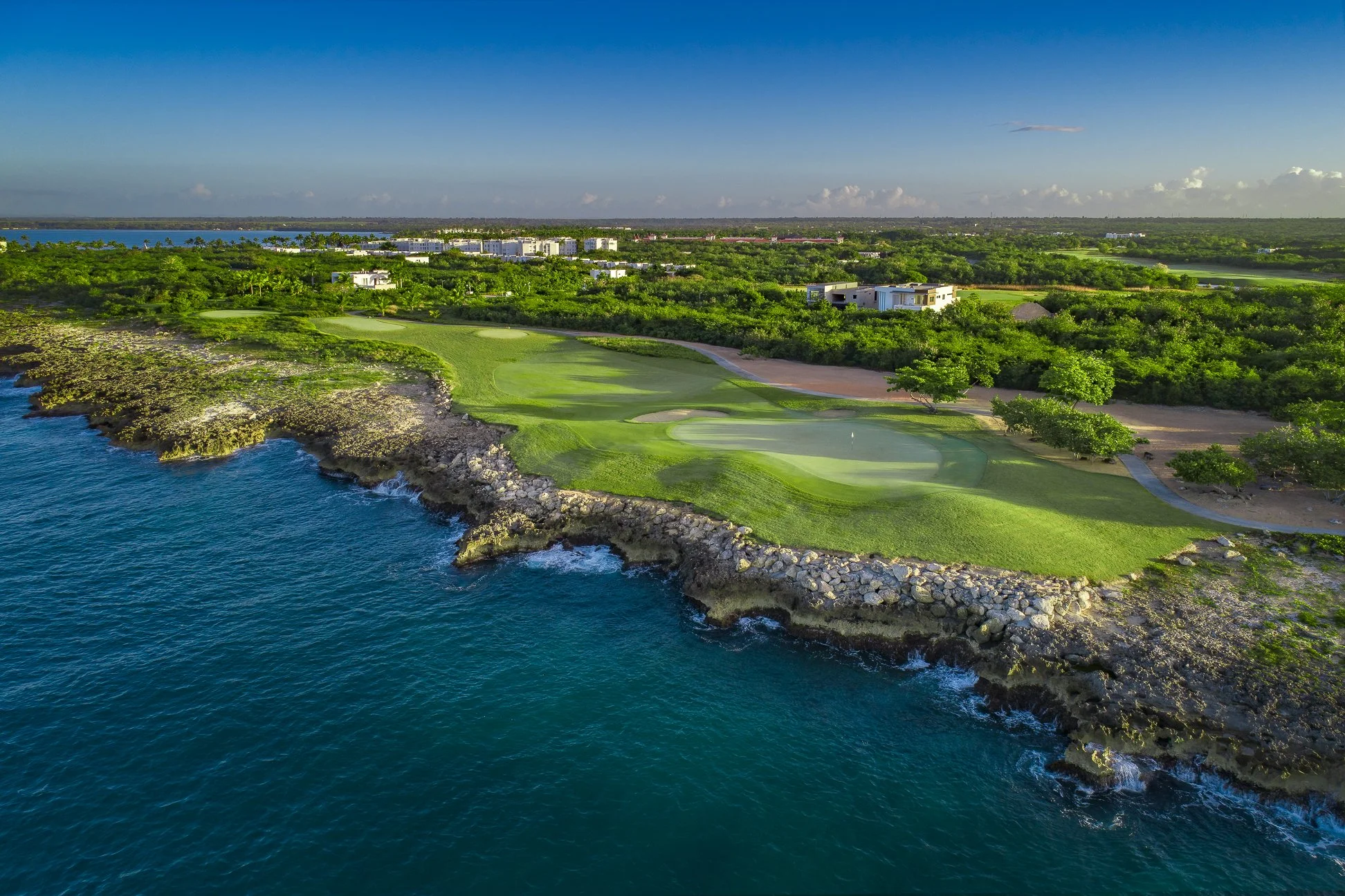A coastal golf course with green fairways and sand traps along a rocky shoreline, overlooking the ocean with blue water and a distant cityscape under a partly cloudy sky.