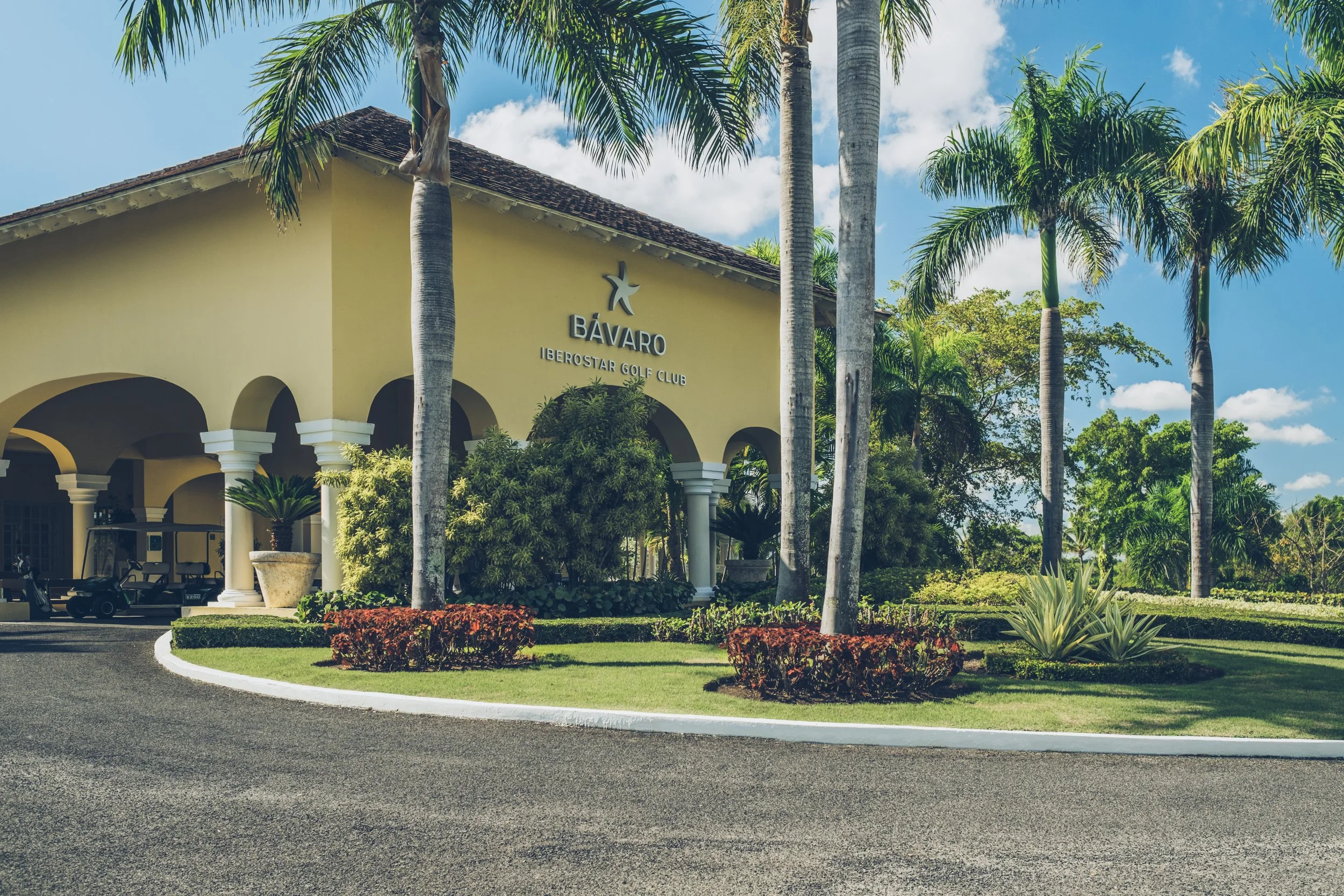 Entrance of Bávaro Iberostar Golf Club surrounded by palm trees, with a yellow building and a parking area, under a blue sky with clouds.