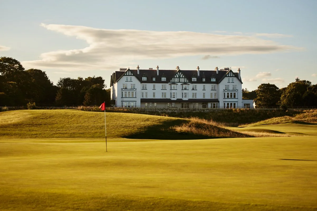 A golf course with a green and a red flag, with a large white mansion-style building in the background under a partly cloudy sky.