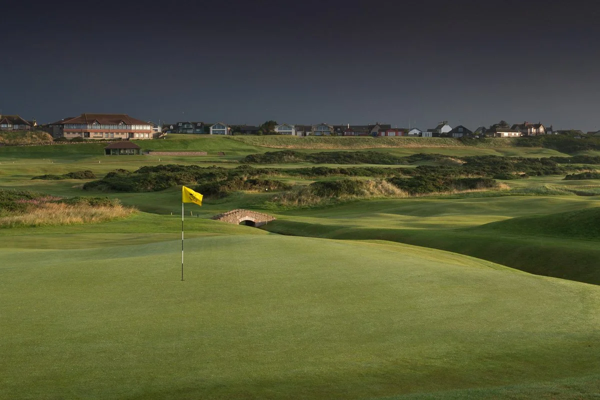 A golf course with a green and flag in the foreground, rolling hills, a small bridge, and houses on a hill in the background under a dark sky.