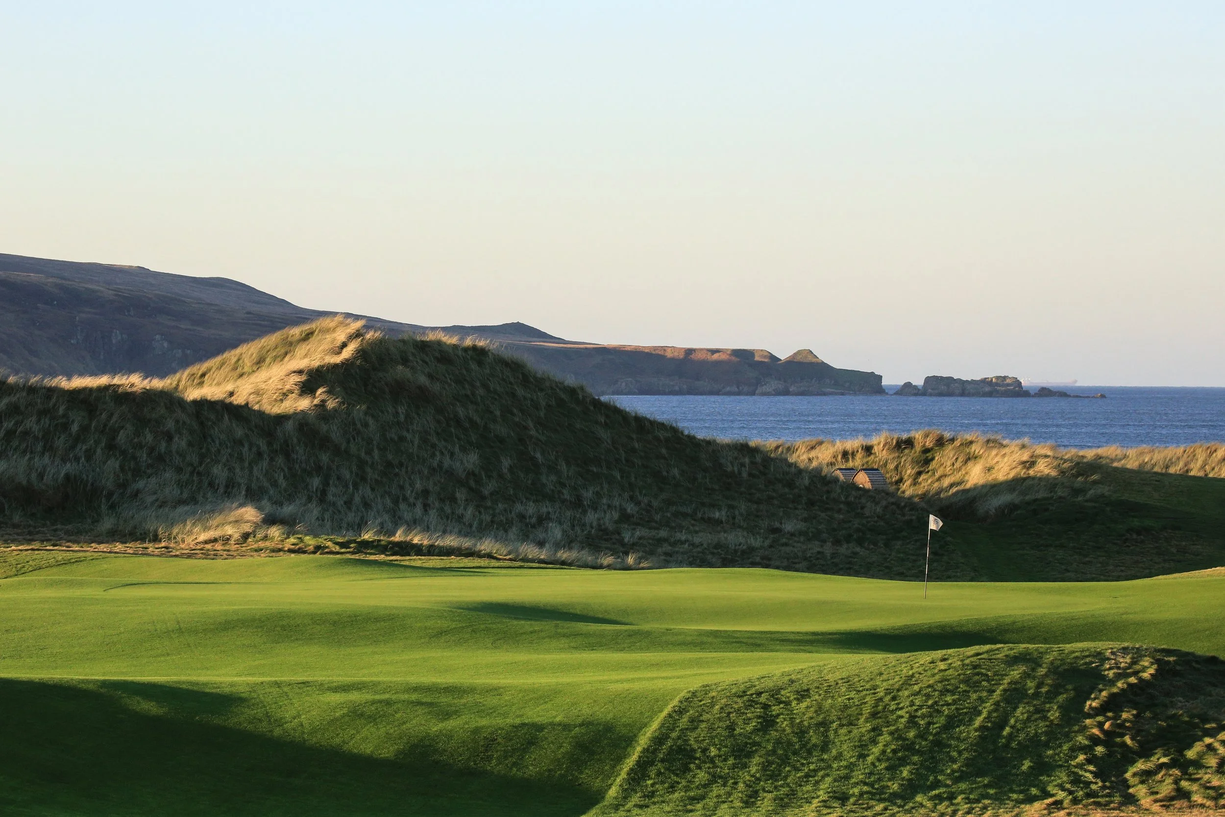 A golf course with green grass, a flag, and sandy dunes, situated along a coastline with cliffs and the ocean in the background during daytime.