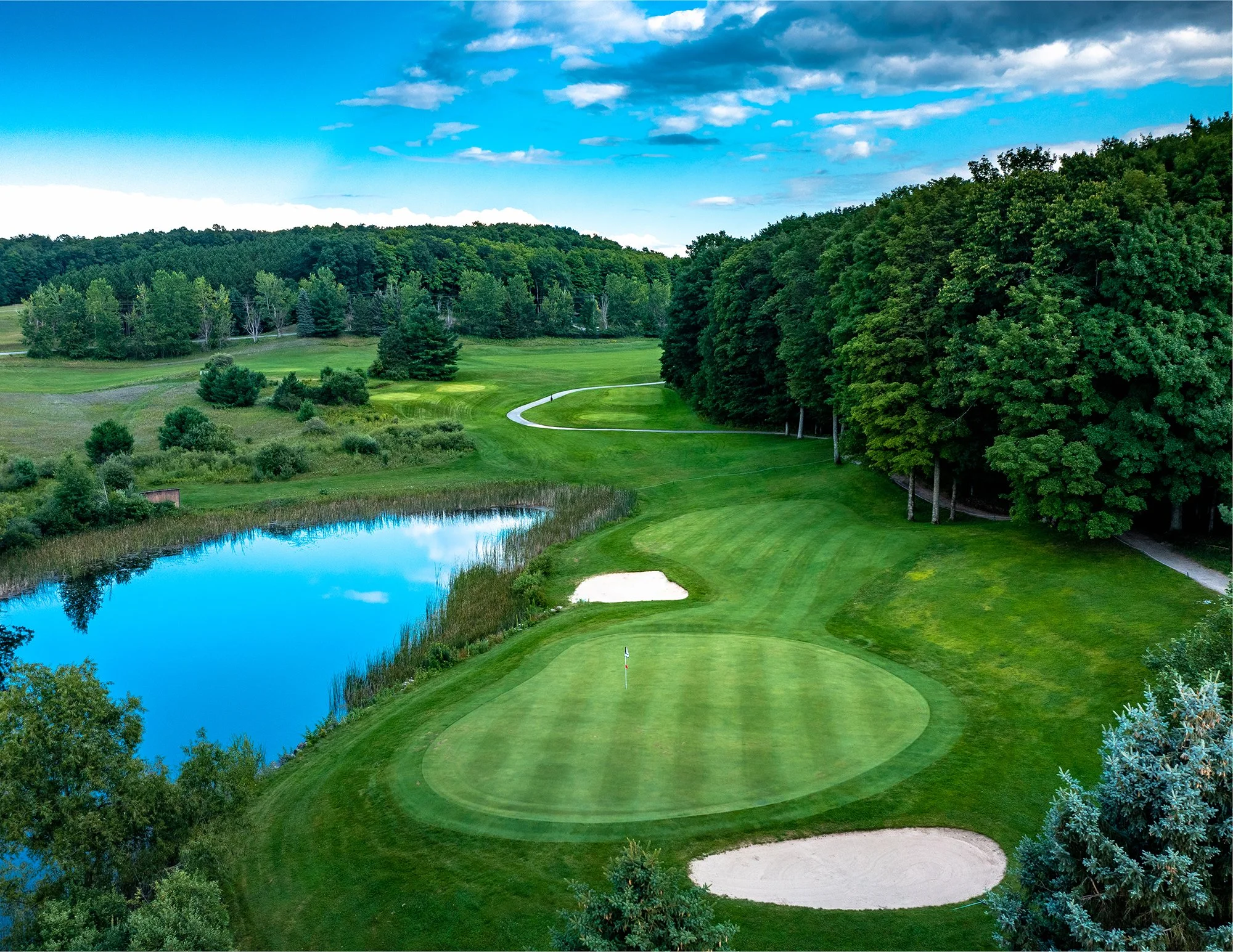 A scenic golf course with a putting green, sand bunkers, a water hazard, and dense trees under a partly cloudy sky.