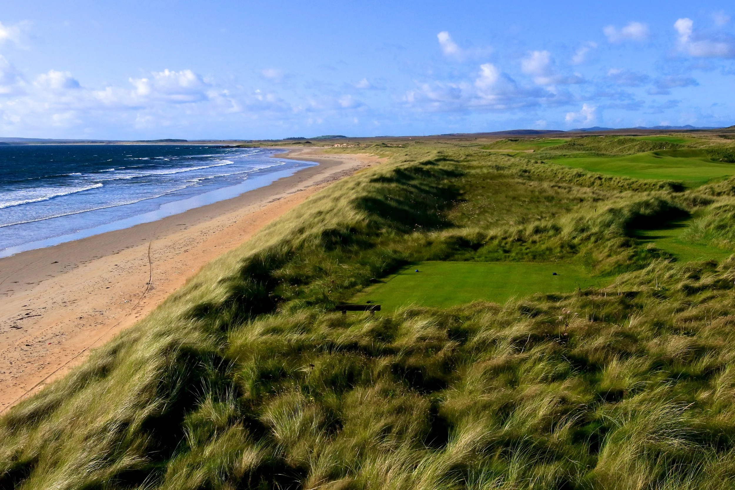 A scenic view of a golf course built into grassy dunes overlooking a beach with ocean waves and blue sky with clouds.