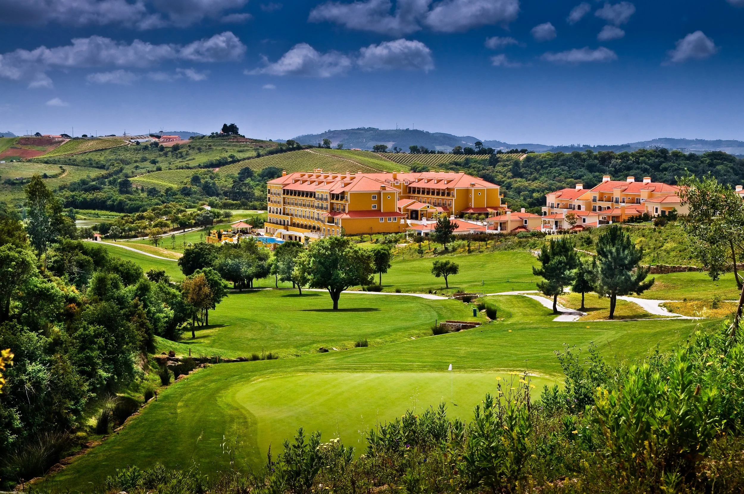 A scenic landscape featuring a large yellow resort building with red roof tiles, surrounded by green trees and grass, with rolling hills and vineyards in the background under a partly cloudy blue sky.