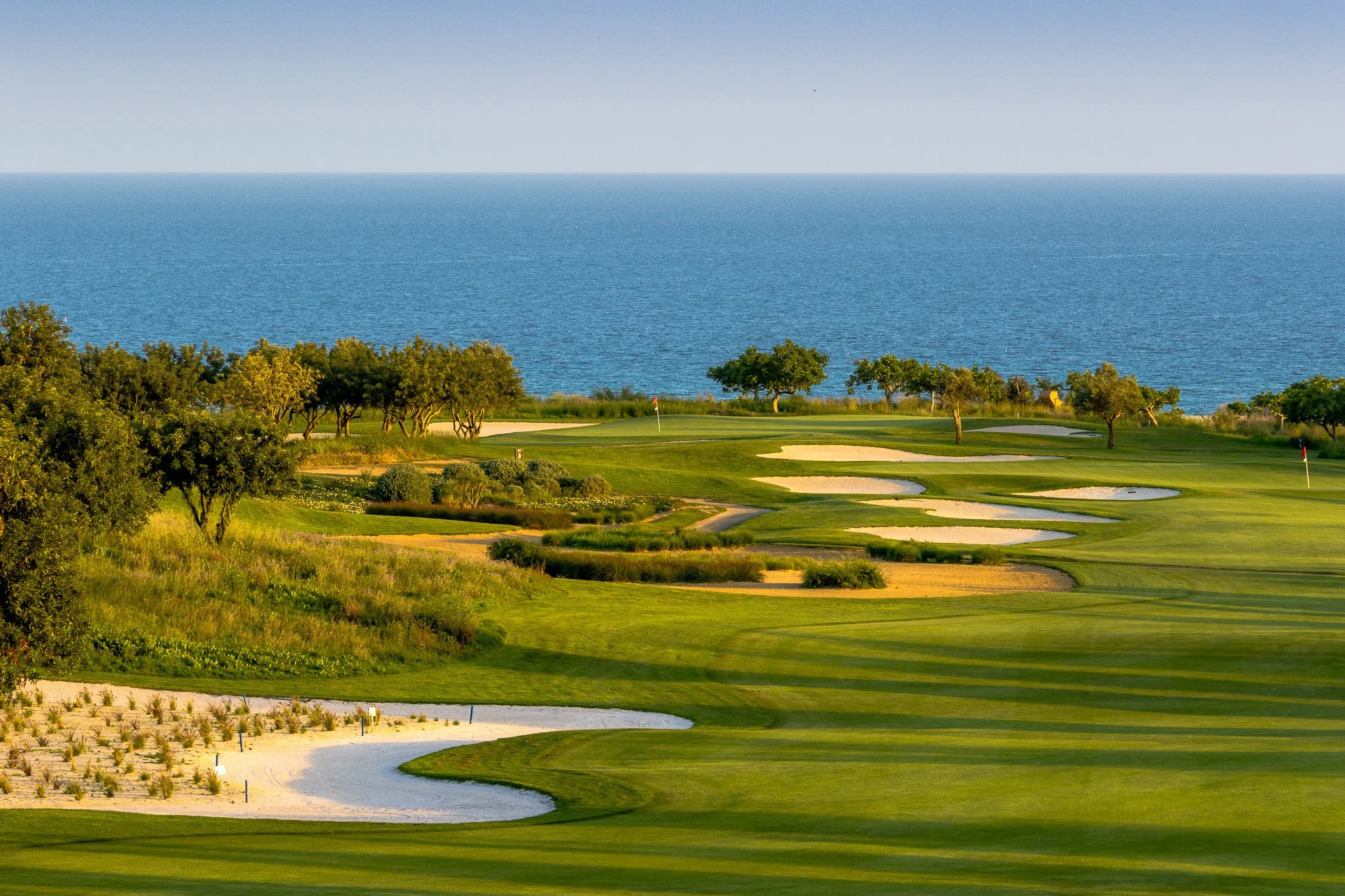 A golf course with green fairways, sand bunkers, and trees near the coast with a large body of water in the background.