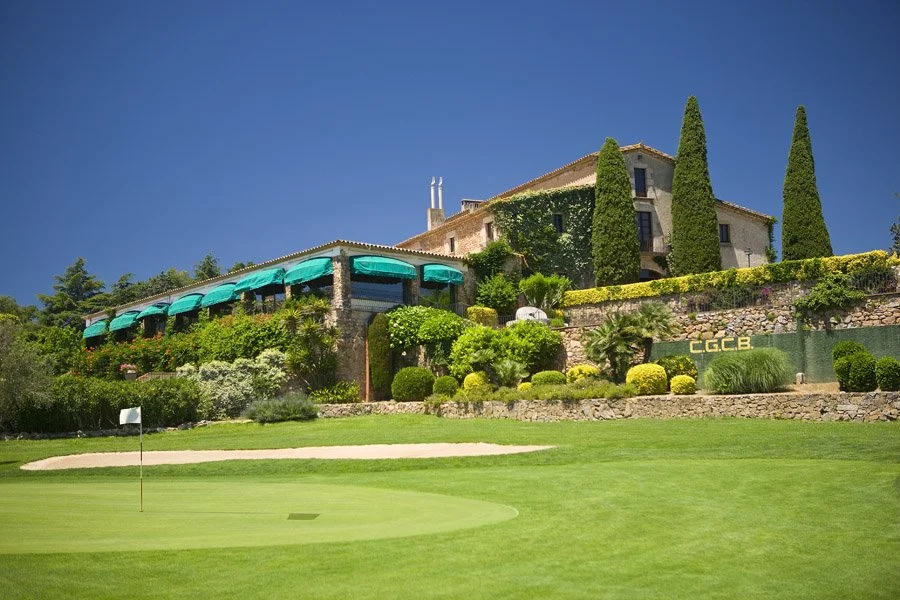 Golf course with a putting green and a sand trap, lush landscaping, and a large house with green awnings and tall cypress trees in the background at a sunny day.