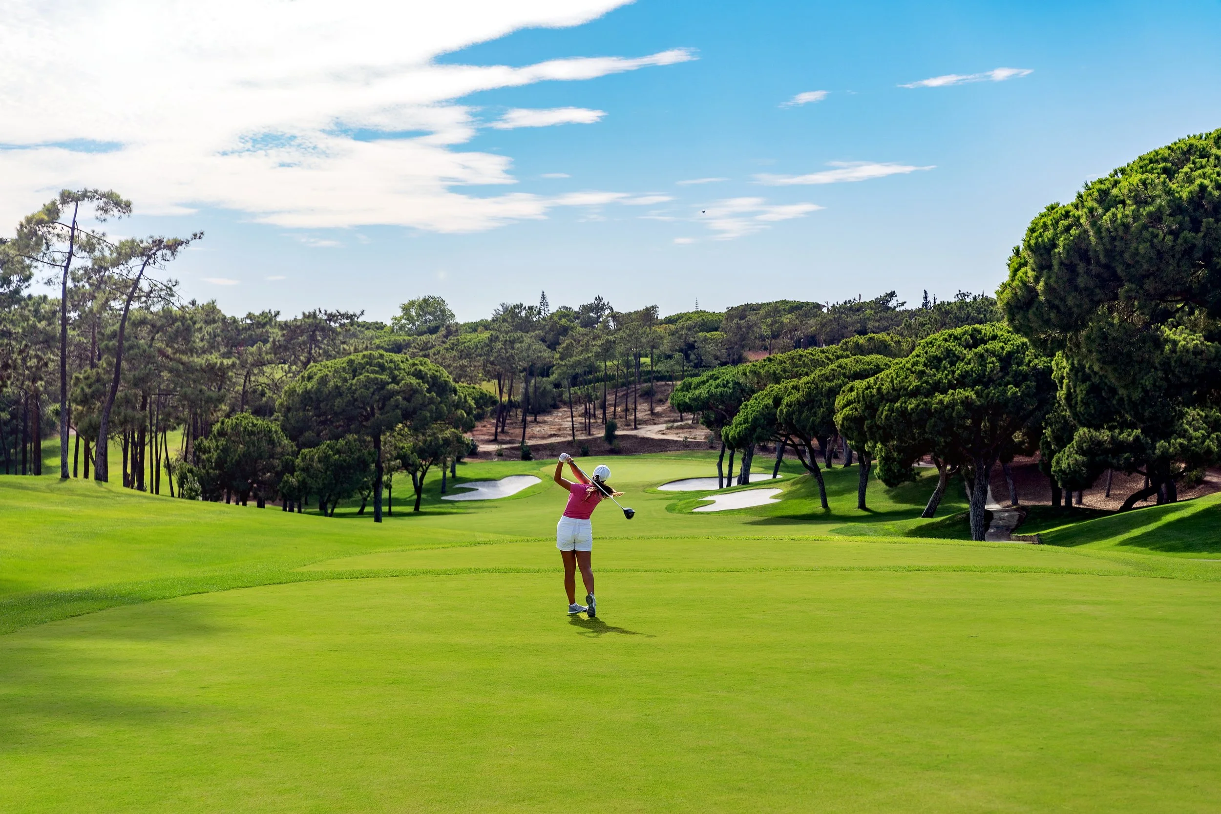 A scenic view of a golf course with green fairways and palm trees beside a body of water, with mountains and a partly cloudy sky in the background.