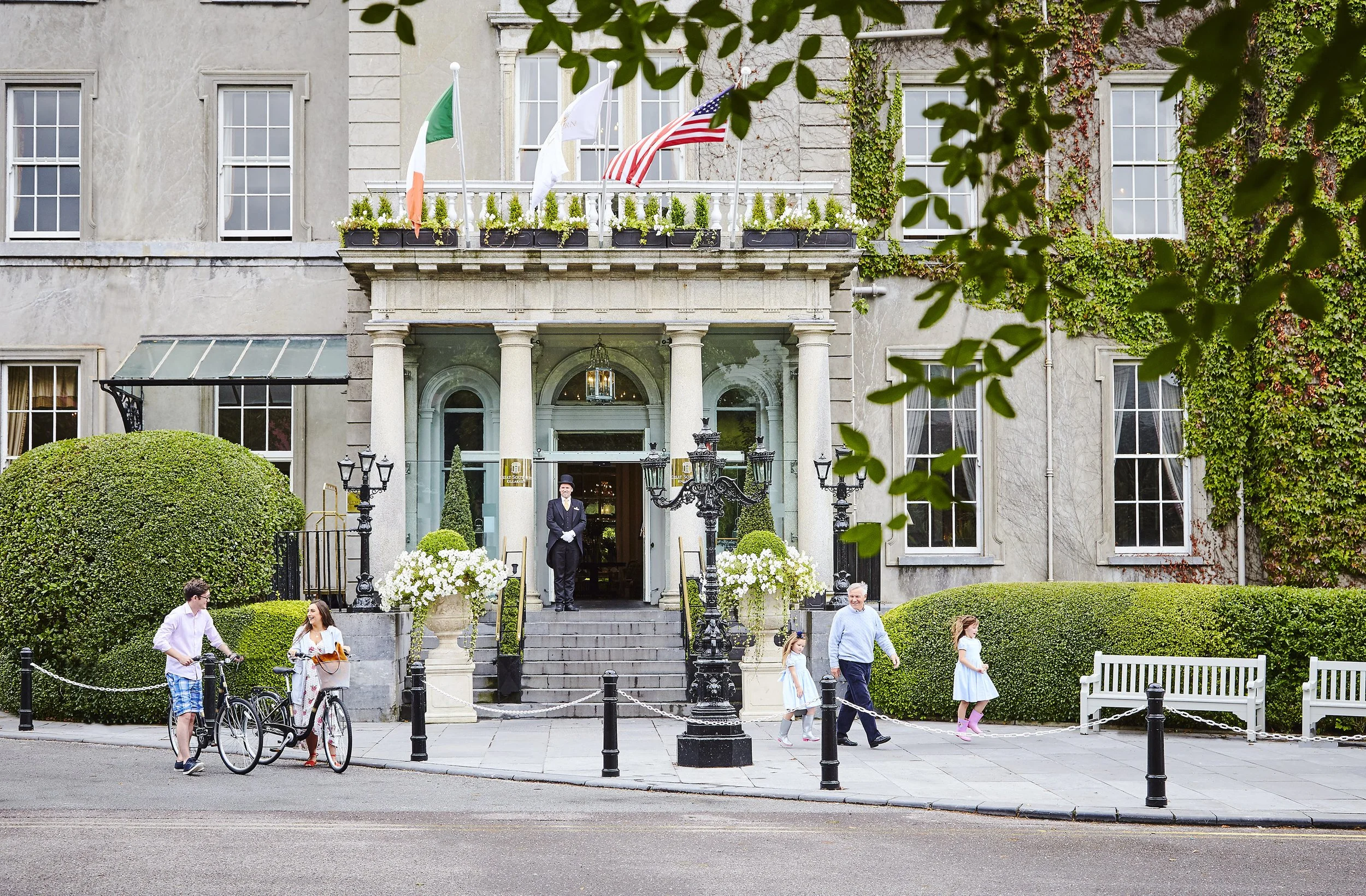 Exterior of a historic hotel with flags, greenery, and people. A doorman stands at the entrance. A man and woman with bicycles and a family walking by.