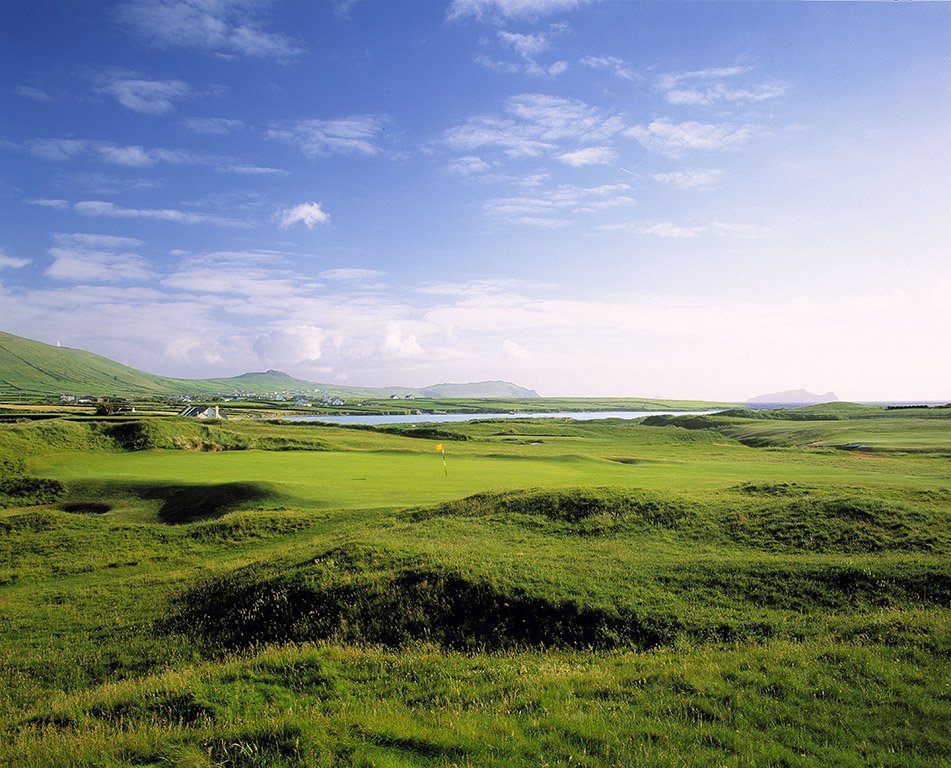 A scenic view of a golf course with well-maintained green grass, distant hills, a water body, and a partly cloudy sky.