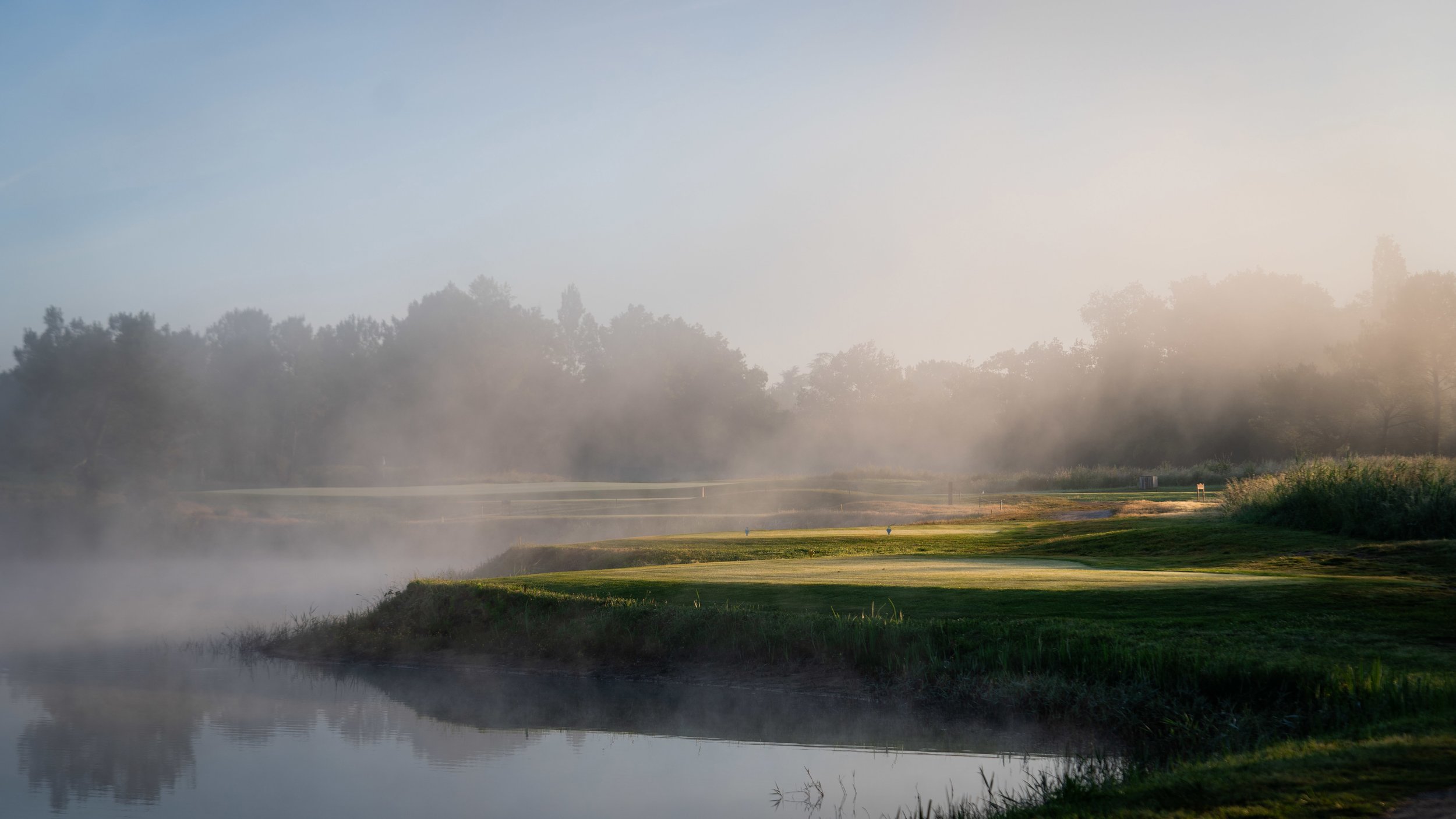 A serene, misty morning view of a lush green fairway alongside a calm pond at Cabot Bordeaux, a premier destination for our luxury golf and wine vacations in Southwest France.