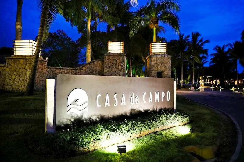 Nighttime view of the entrance sign for Casa de Campo, illuminated lights, palm trees, stone pillars, and a curved road.