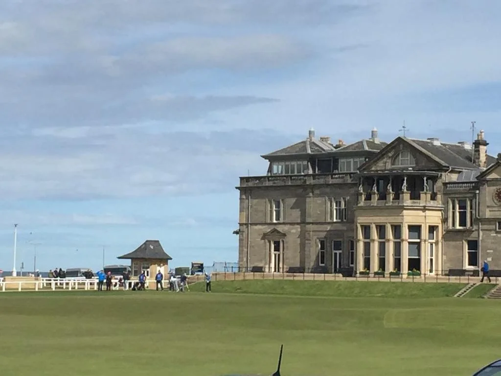 A large historic building with a castle-like appearance located near a golf course, with several people and vehicles around, under a partly cloudy sky.