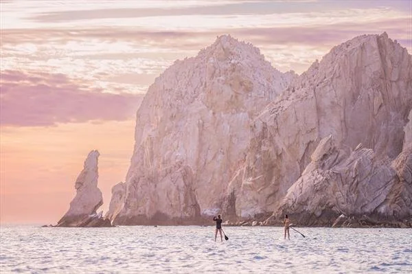 Two people stand paddleboarding in front of large rocky cliffs during a sunset with pink and purple sky.