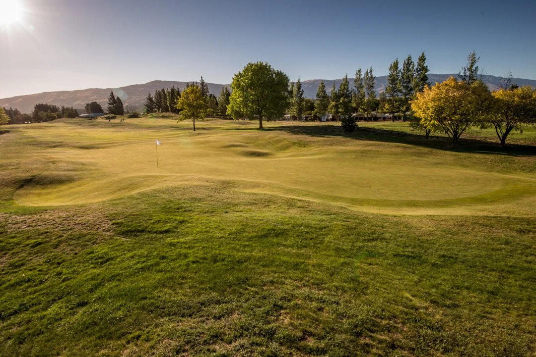 A golf course with a putting green, a flagstick, and trees, with mountains in the background and a clear blue sky.