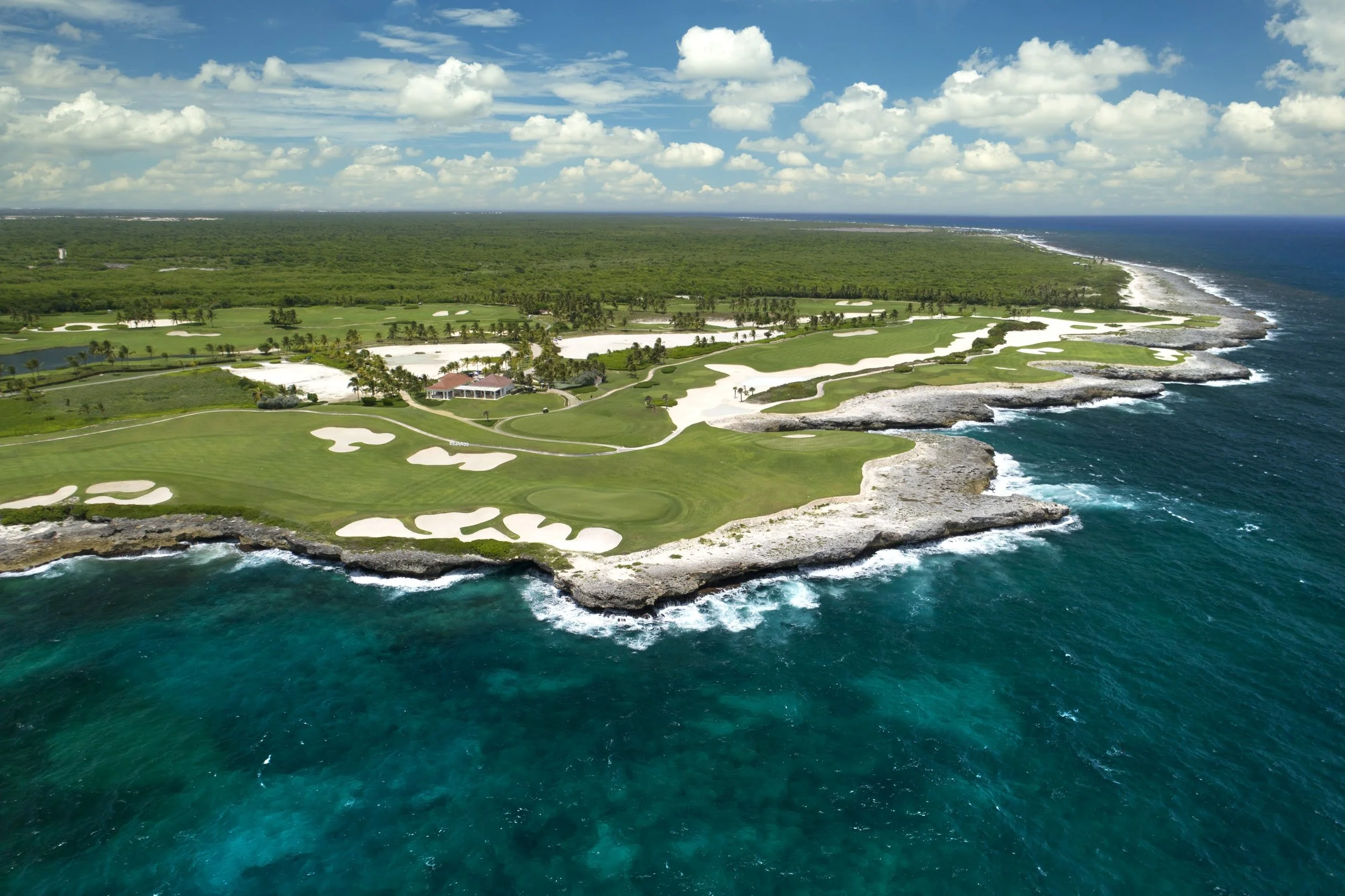 Aerial view of a coastal golf course with green fairways, white sand bunkers, and rocky shoreline along the ocean under a partly cloudy sky.