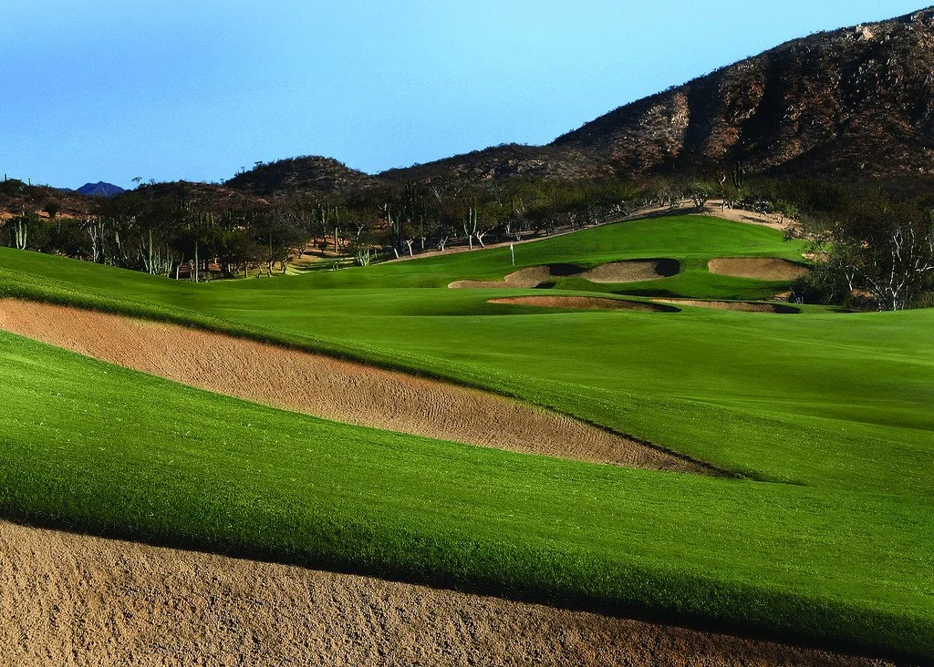 A scenic view of a golf course with lush green fairways, sand bunkers, and a backdrop of mountains and trees under a clear blue sky.