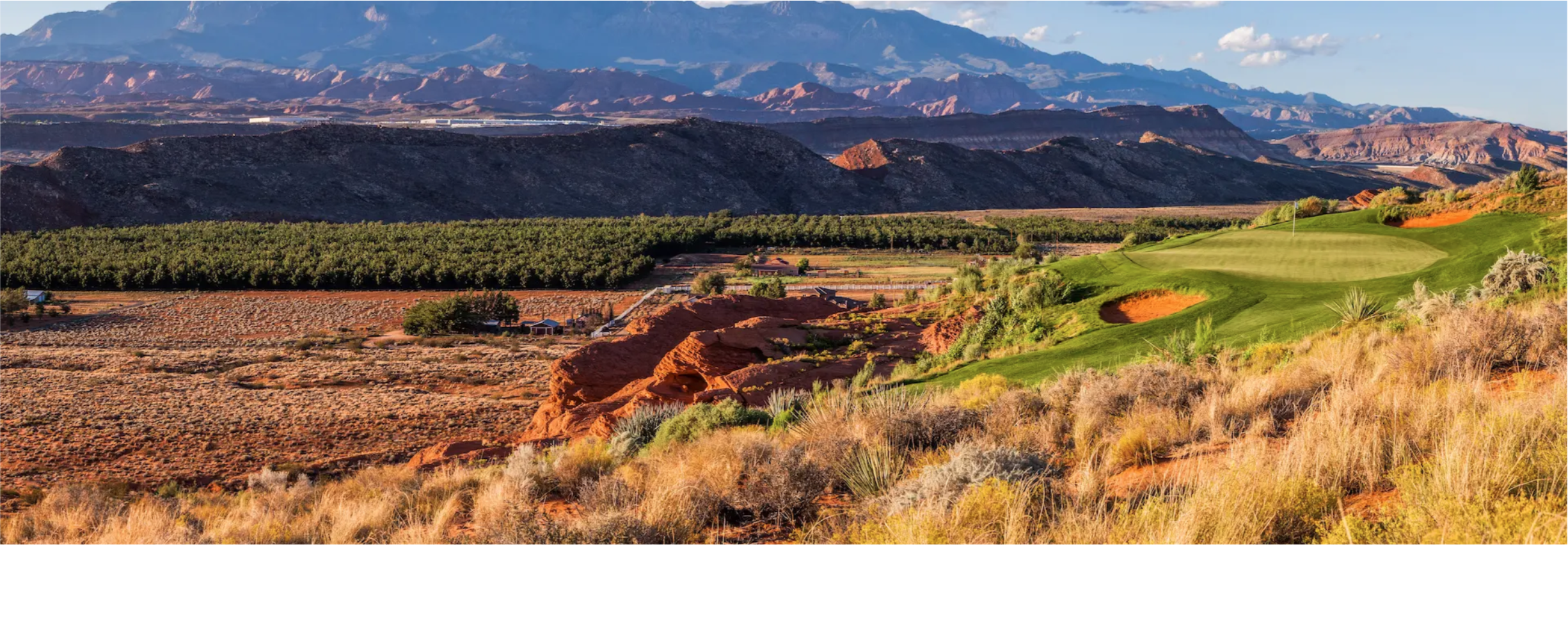 Scenic landscape with mountains in the background, a golf course with green fairways, and desert terrain with dry grasses and shrubs in the foreground.