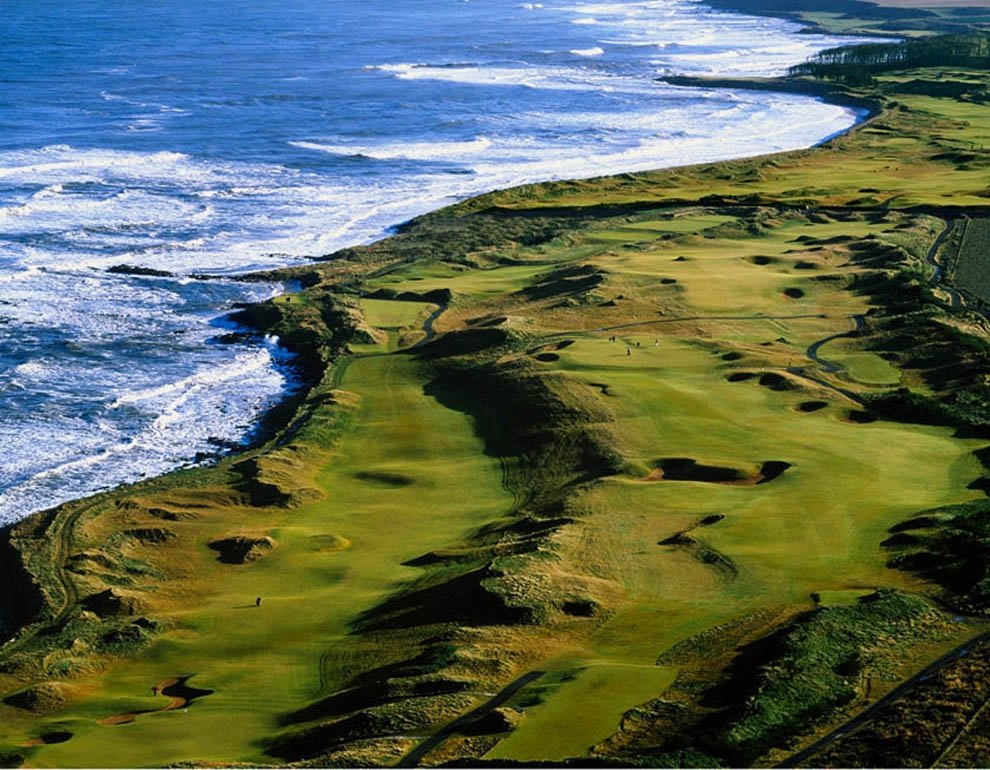 Aerial view of a coastal golf course with lush green fairways and multiple sand bunkers, bordered by ocean waves on one side and rolling hills on the other.