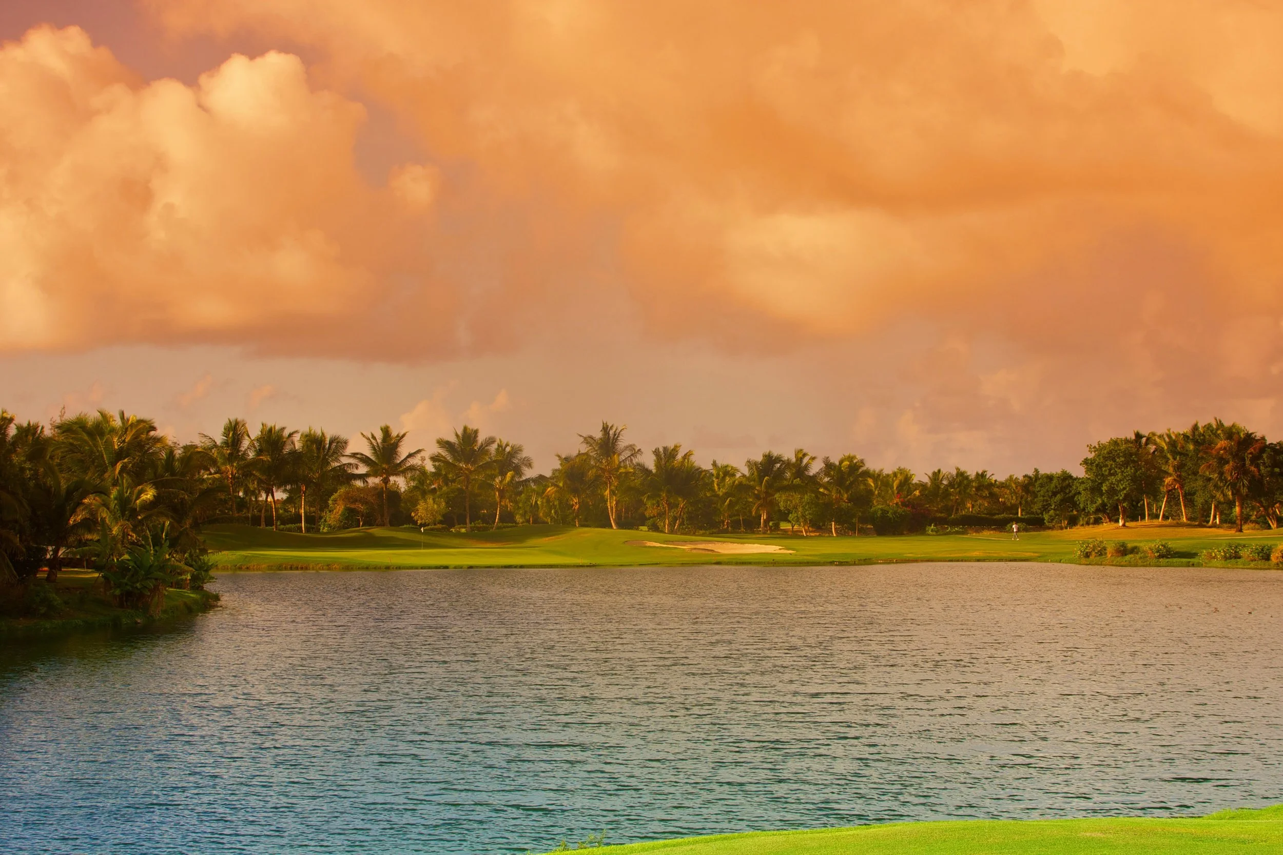 Golf course with water, green grass, and palm trees under a partly cloudy sky at sunset.