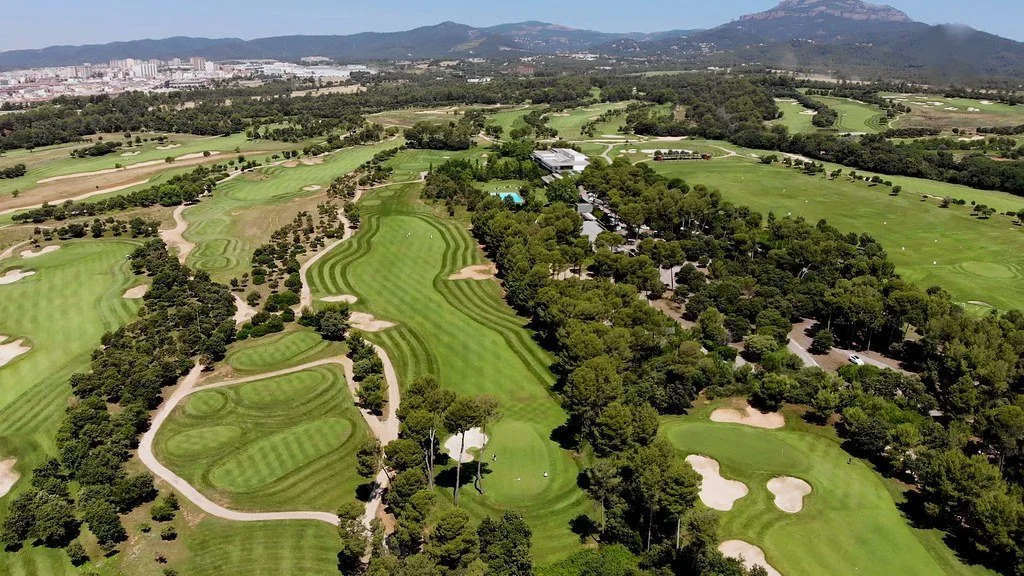 Aerial view of a large golf course with lush green fairways, sand bunkers, and numerous trees, with mountains and a city in the background.