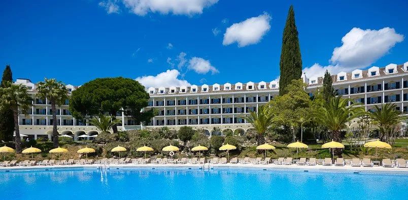 Resort hotel with a swimming pool, yellow umbrellas, and tropical trees under a blue sky with clouds.