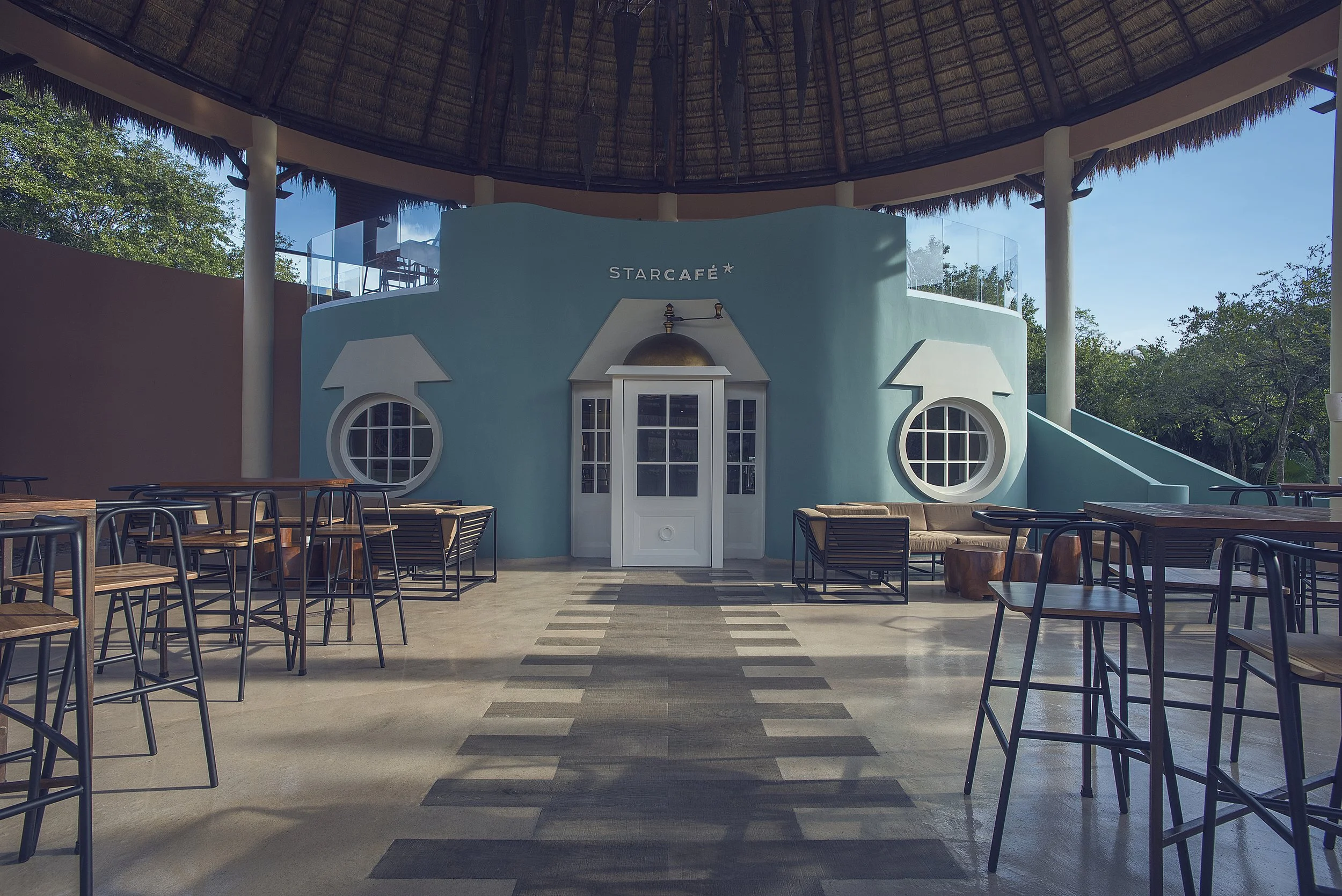 Interior of a tropical-themed cafe with pastel blue walls, round window accents, and outdoor seating with wooden chairs and tables, under a thatched roof.