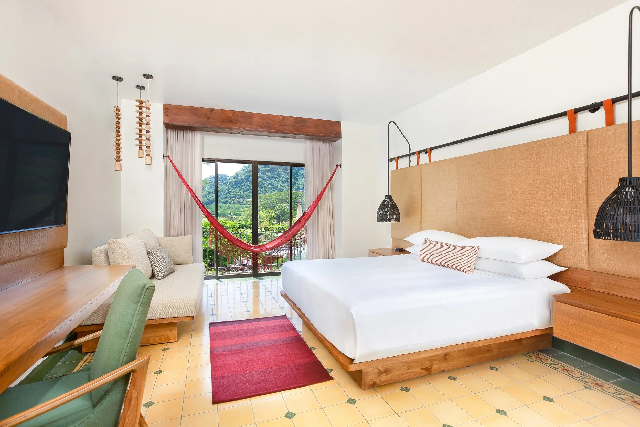 Hotel room with a large bed, wooden headboard, black hanging lamps, green tiled floor, beige sofa, wooden desk, and a sliding glass door with a view of greenery and a red hammock outside.