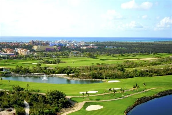 A scenic view of a golf course with green fairways, sand traps, and water hazards, overlooking a cityscape and the ocean in the distance under a partly cloudy sky.
