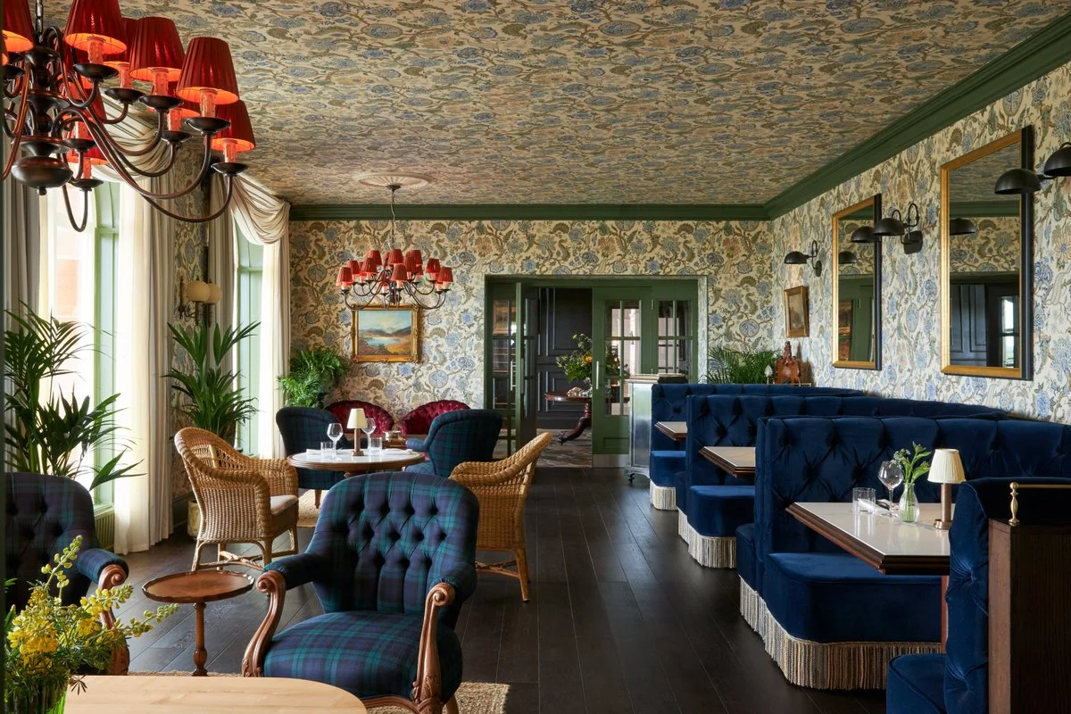 A vintage-style interior with patterned wallpaper, green trim, dark wooden floors, lush greenery, wicker and upholstered chairs, blue velvet booth seating, framed artwork, lamps, and red lampshade chandeliers.