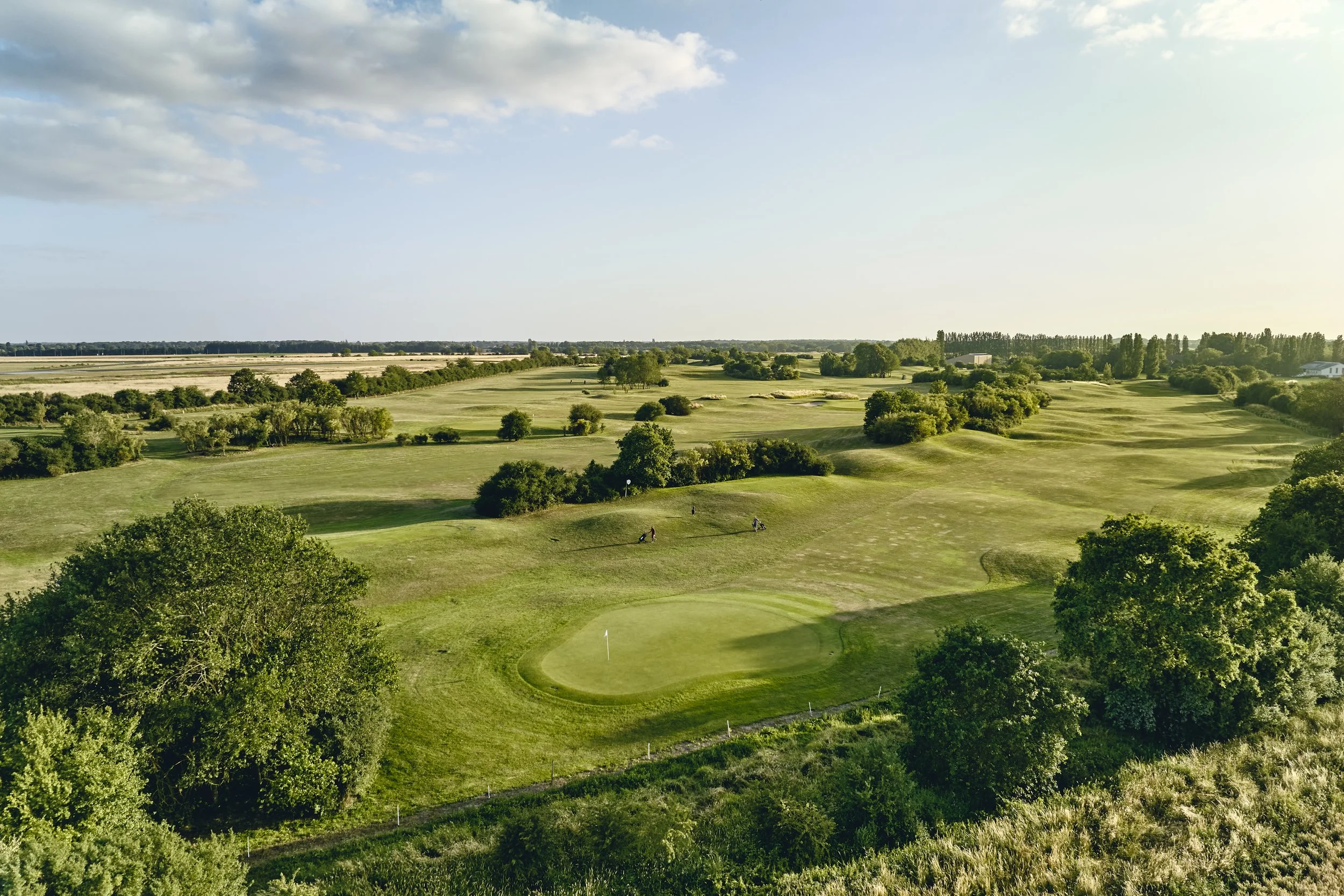 Aerial view of a golf course with green fairways, sand bunkers, and trees, under a partly cloudy sky.