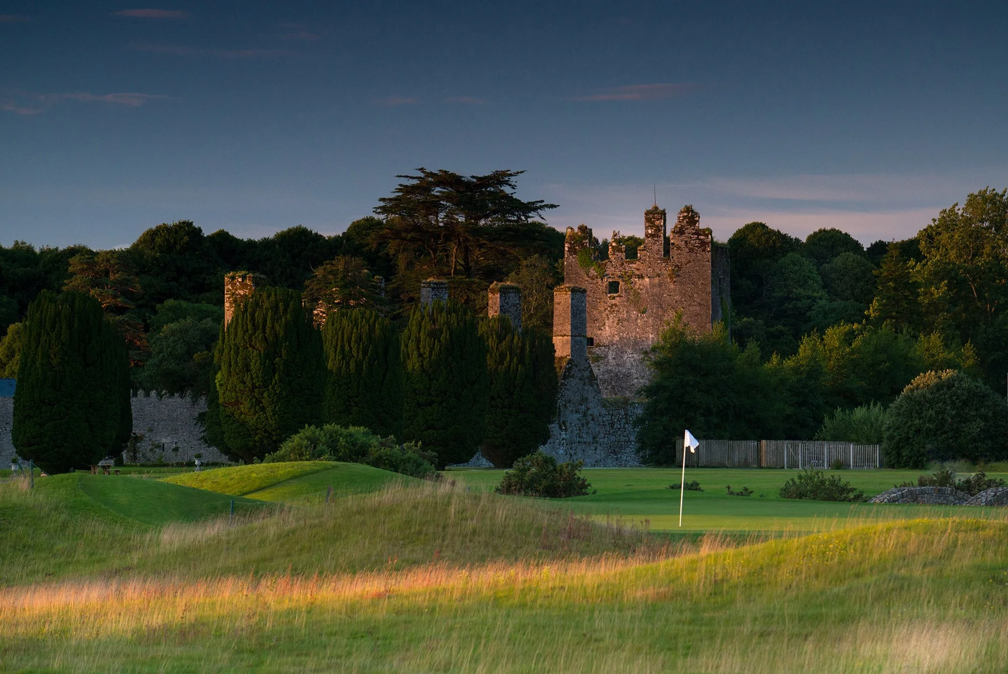 A castle on a hilltop surrounded by trees, with a golf course and a flag in the foreground at sunset.