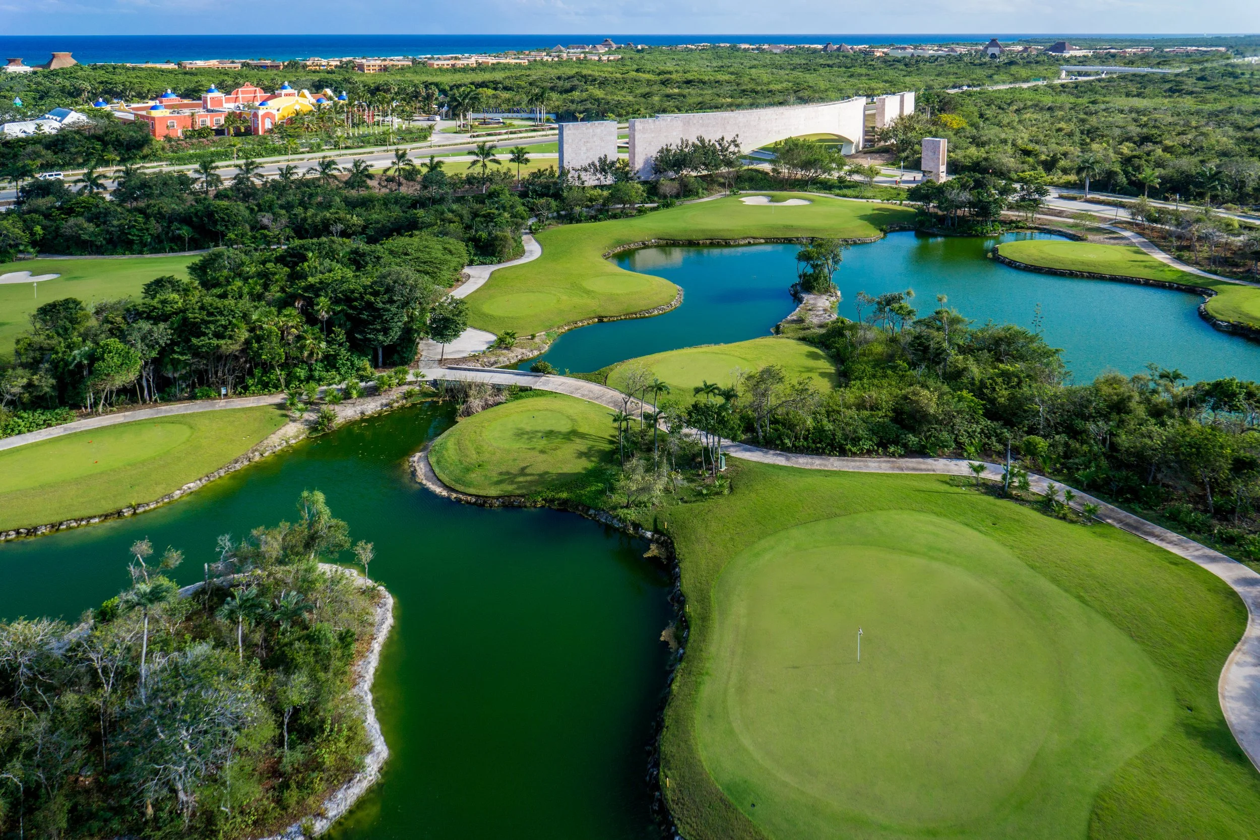 Aerial view of a golf course with green fairways, water hazards, and a putting green with a flag. In the background, there are trees, a road, a colorful building, and the ocean.