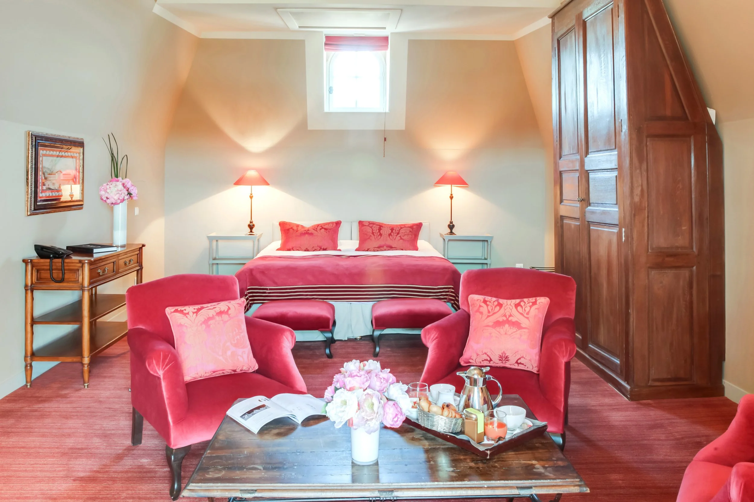 A cozy bedroom with a double bed flanked by two nightstands with lamps, a window above the bed, a wooden wardrobe on the right, and two red armchairs with matching pillows facing a wooden coffee table with a flower arrangement and breakfast items, all on a reddish carpet.