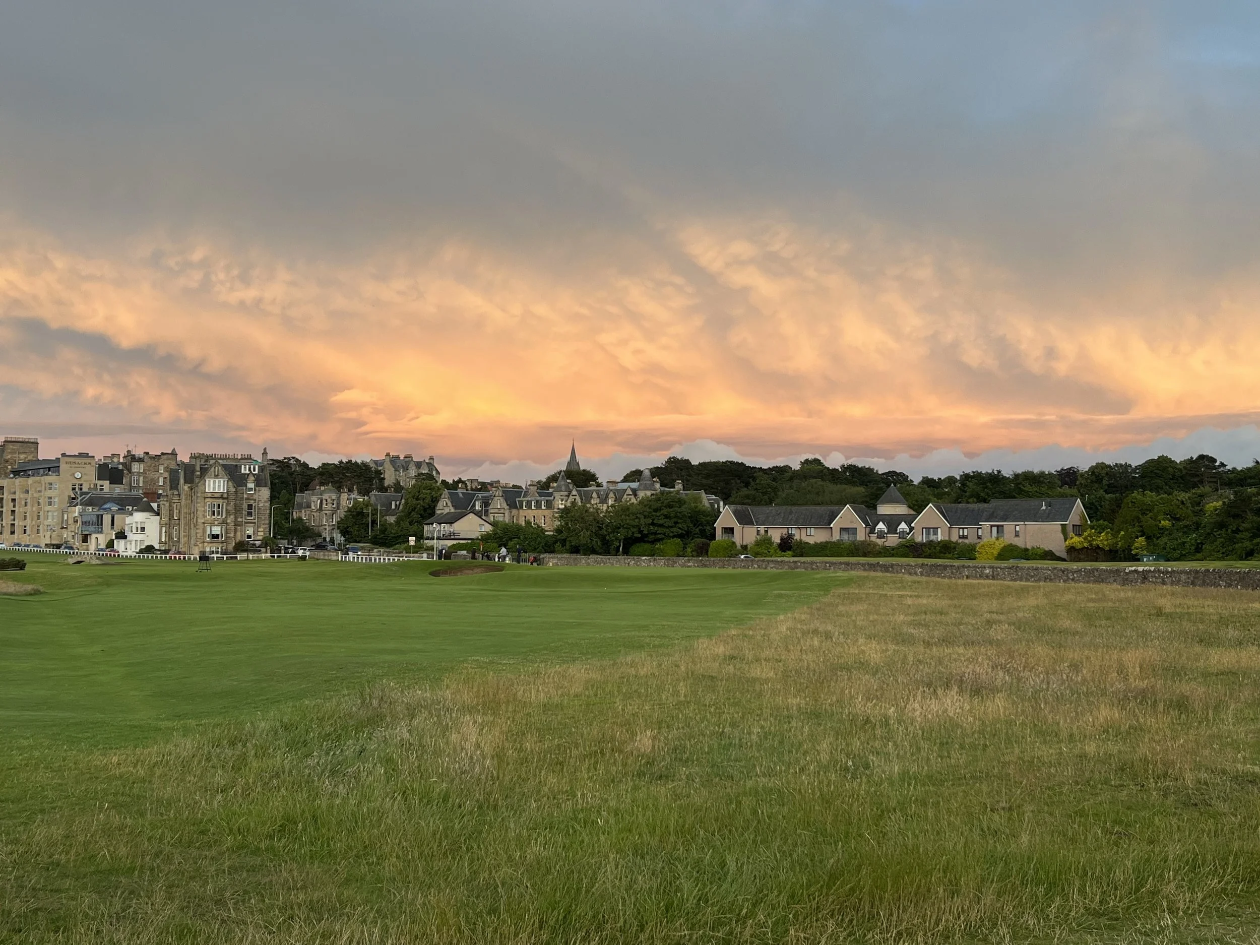 A stunning view from the tall grass of the right-hand rough on the famous 17th hole at the St Andrews Old Course in Scotland, looking toward the fairway and historic town buildings under a dramatic sunset sky.