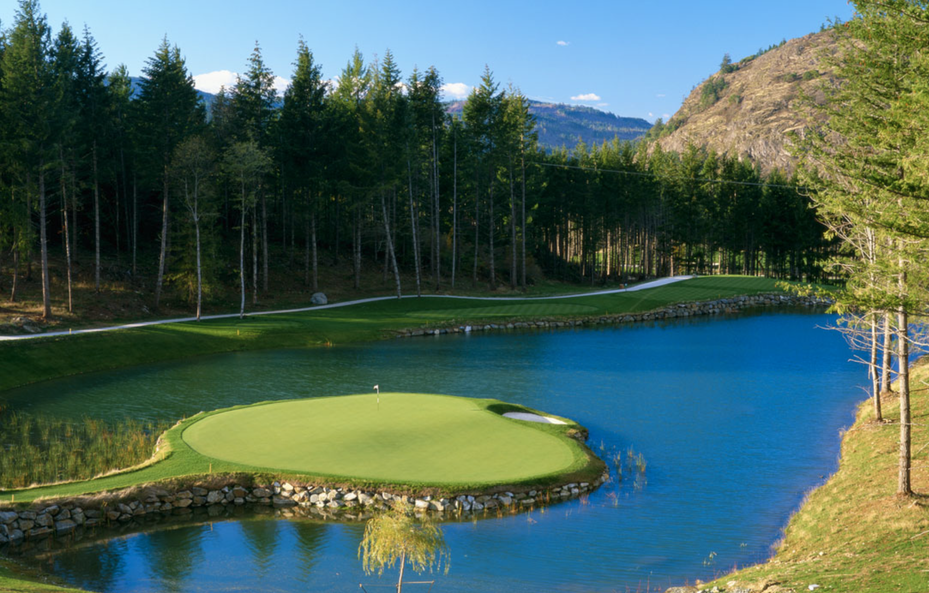 A golf course with a putting green surrounded by water and trees, with mountain and blue sky in the background.