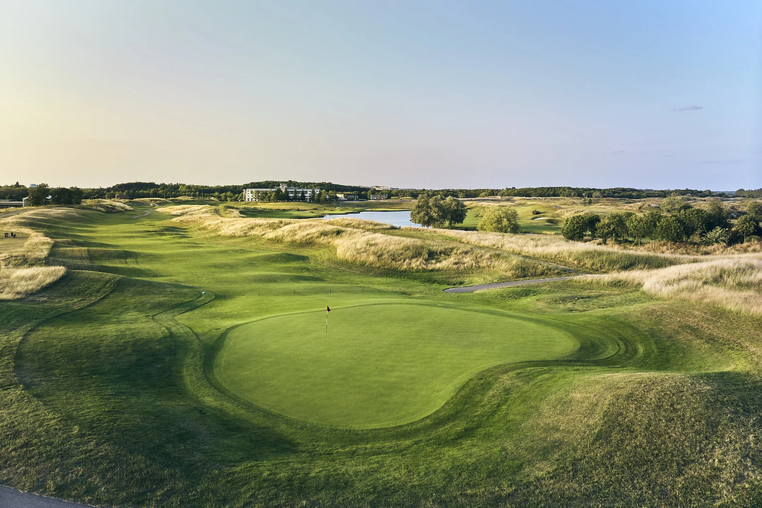 A golf course with a green, flag, and sand traps, surrounded by grass, trees, and a water body in the background.