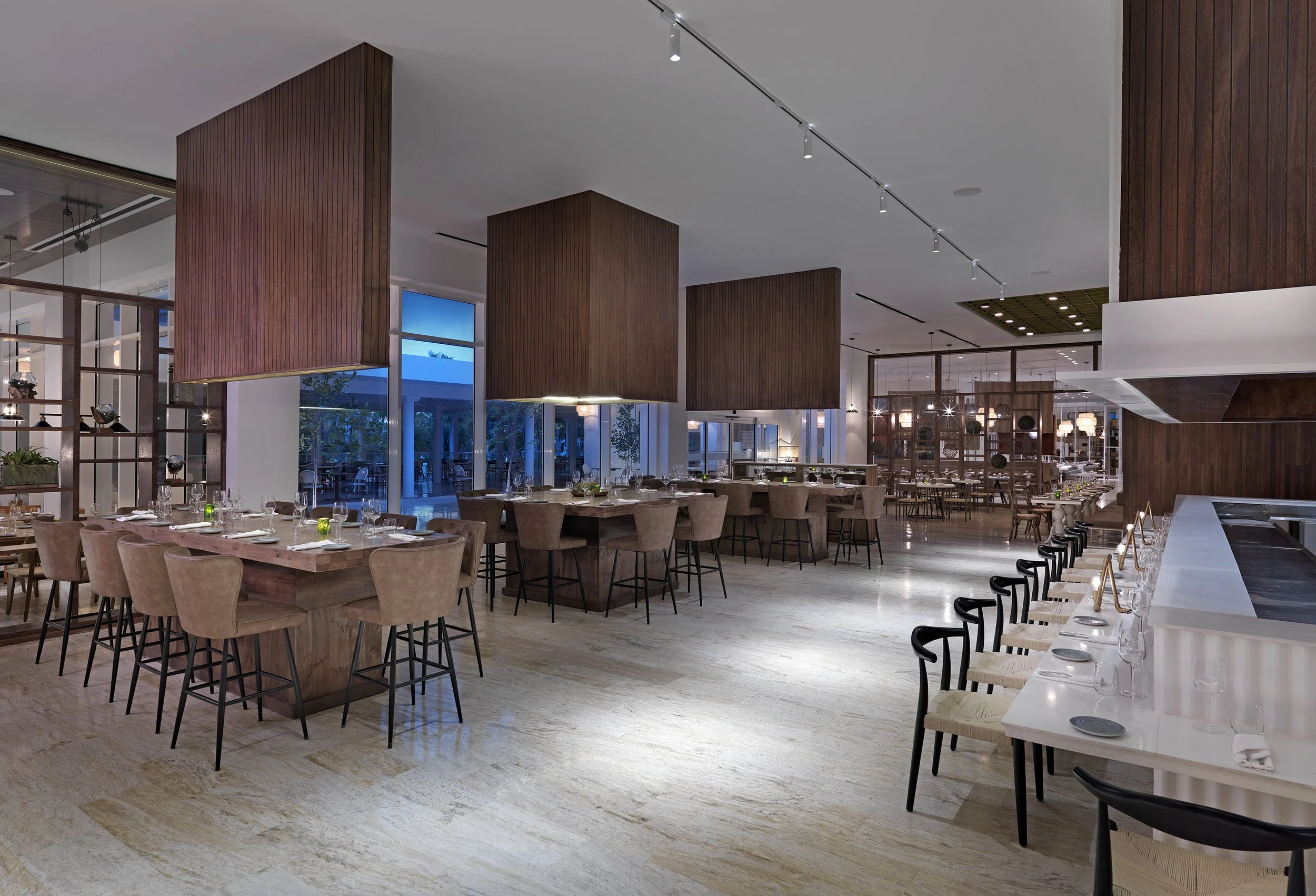Elegant restaurant interior with modern furniture, wooden accents, and large windows showing outdoor seating at dusk.