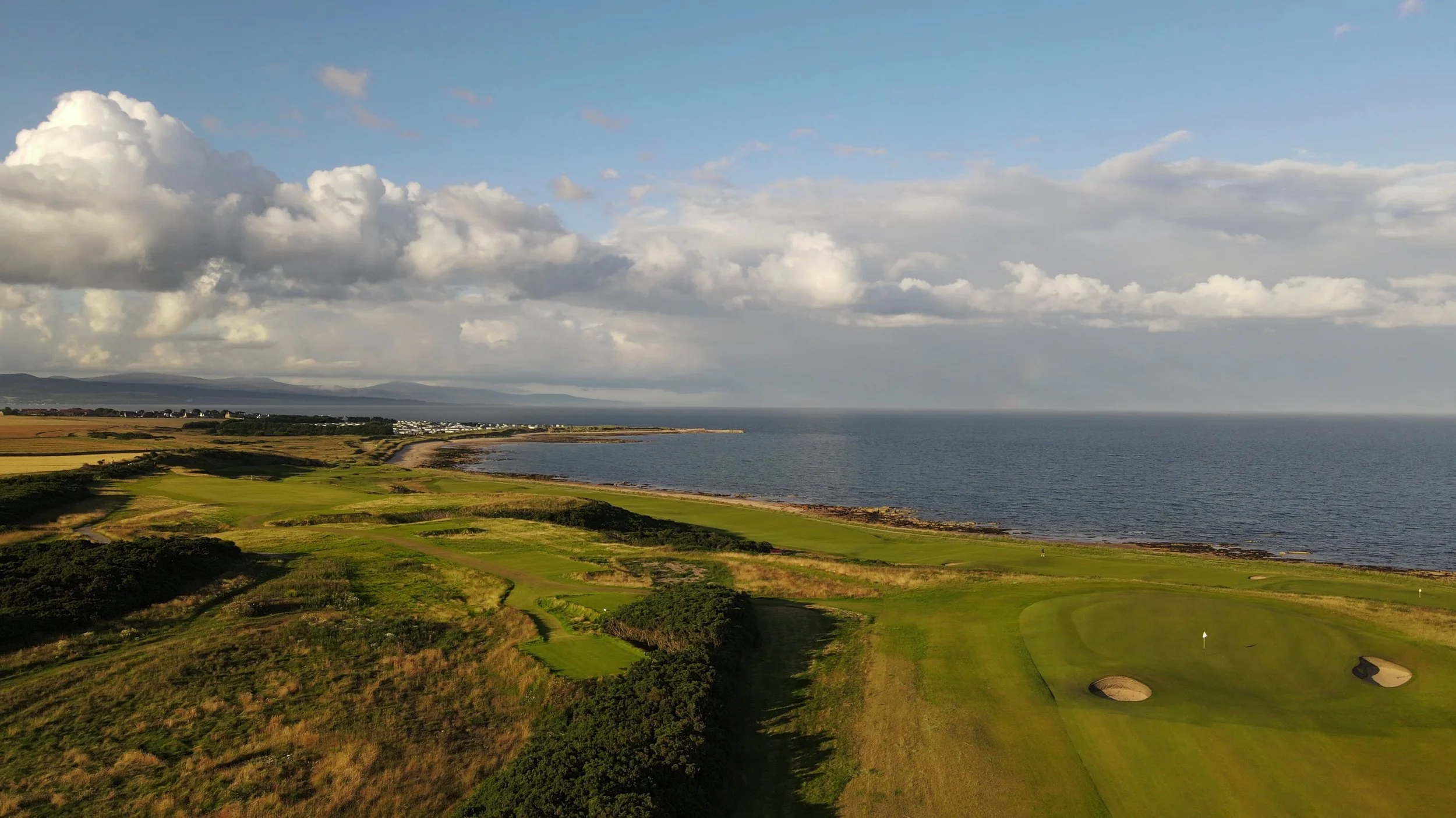 A scenic view of a golf course bordered by the ocean, with green putting greens, sand bunkers, and rolling fairways under a partly cloudy sky.