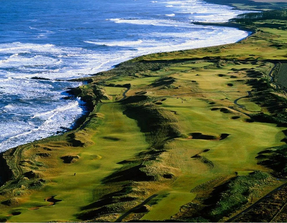 A scenic aerial view of a coastal golf course with green fairways and sand bunkers, adjacent to the ocean's shoreline.