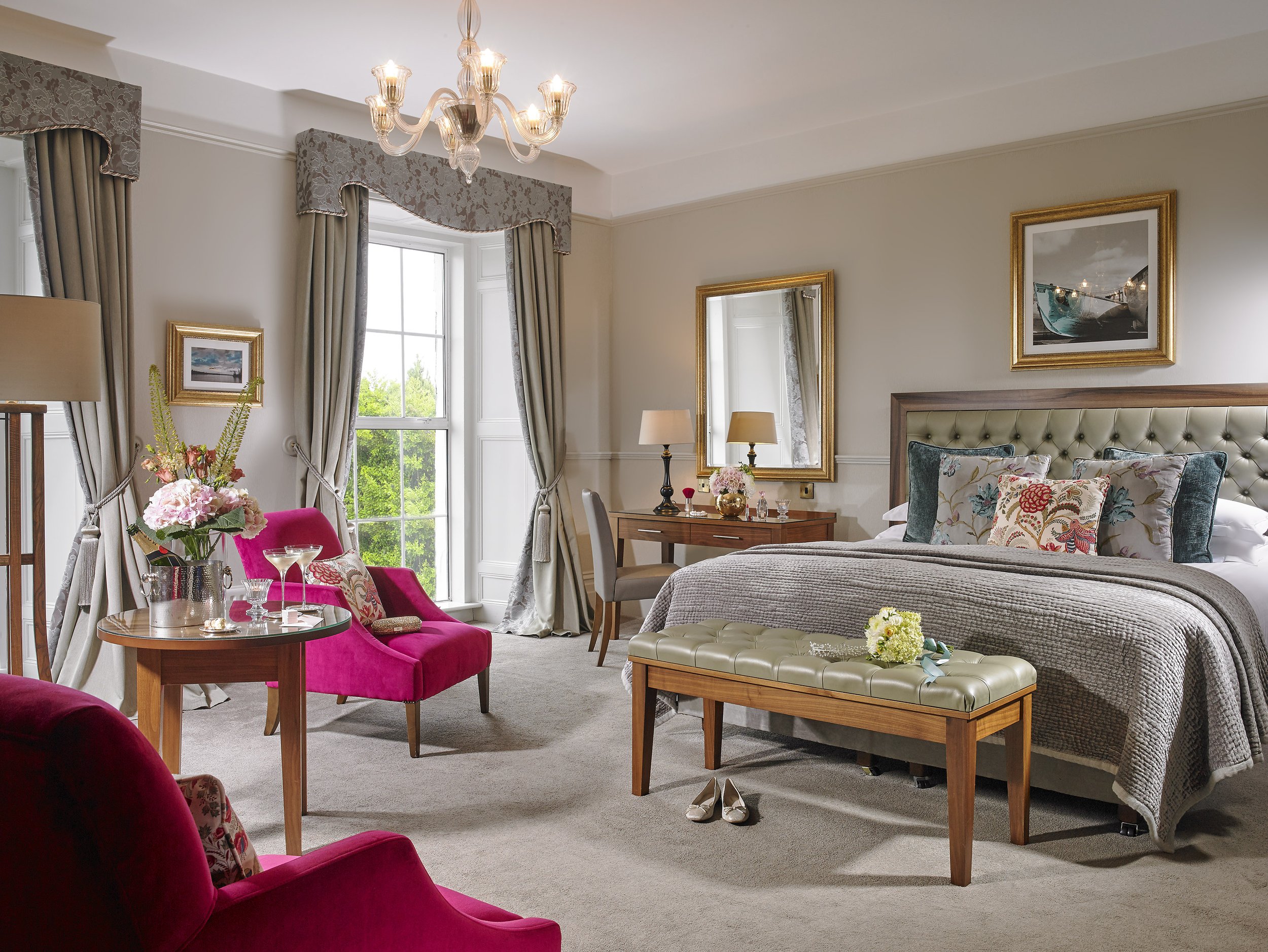 A luxurious bedroom with a large bed, pink chairs, a window with drapes, and elegant decor.
