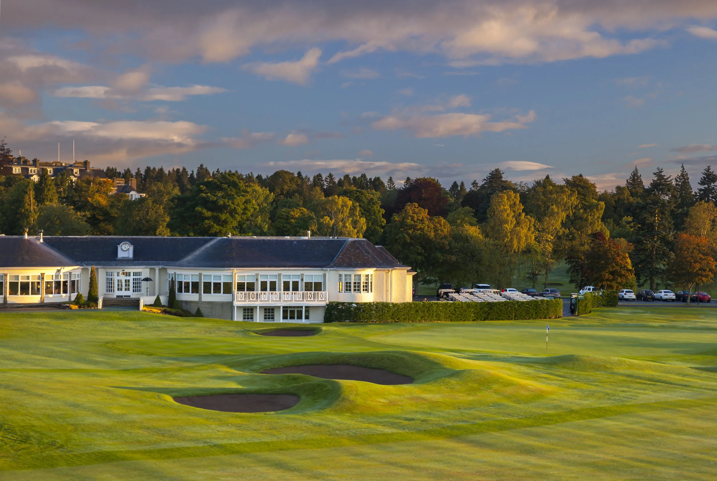 The sweeping 18th green and bunkers at Gleneagles bathed in evening light, with the Dormy clubhouse and the grand Gleneagles Hotel nestled in the trees, a luxurious highlight of our escorted golf tours in Scotland.